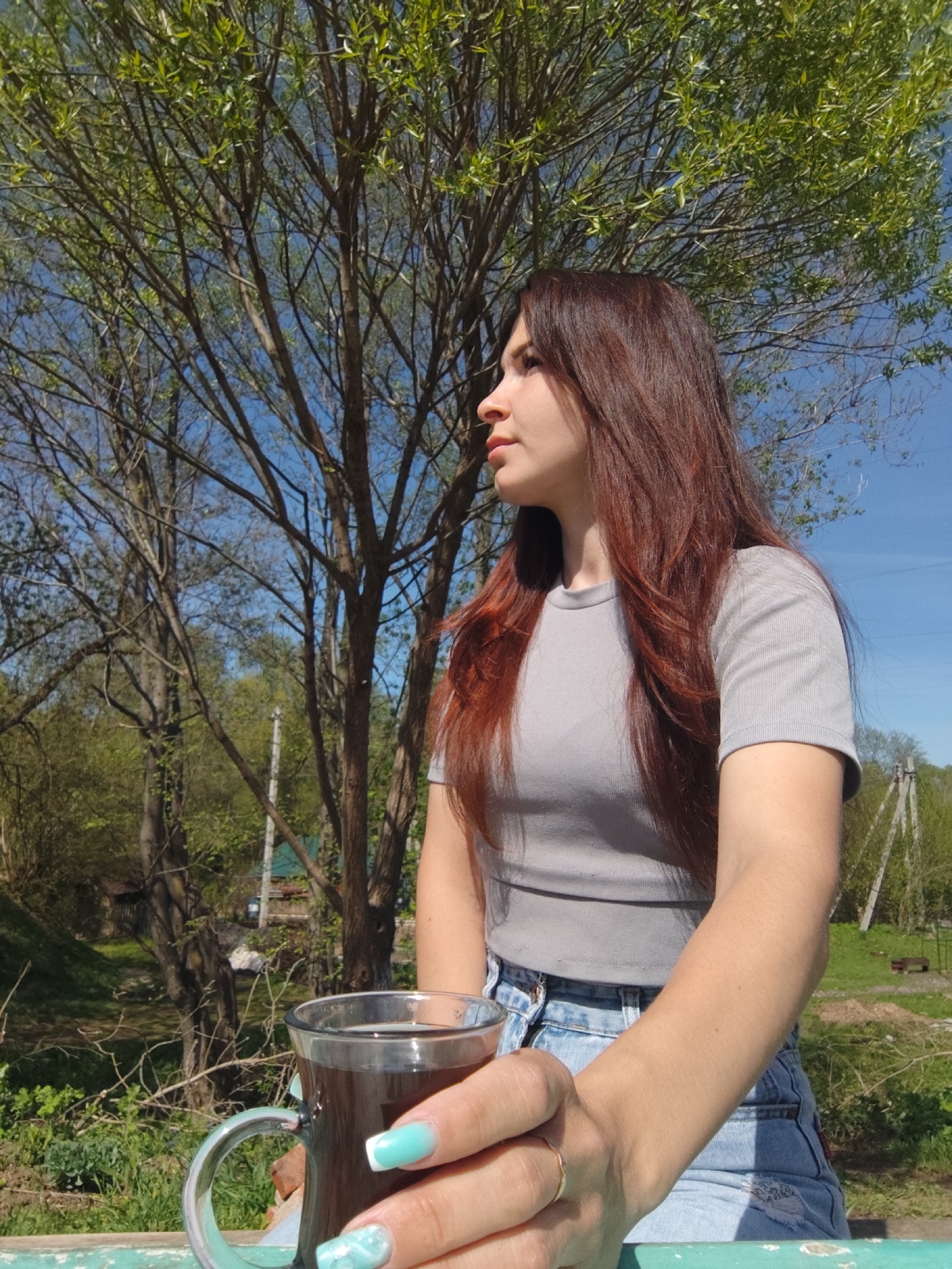 A woman with auburn hair sits outdoors in Фоки, Russia, holding a glass mug of dark liquid. She is wearing a gray t-shirt and light blue denim shorts. The background includes trees with budding leaves, a blue sky, and glimpses of the rural landscape.