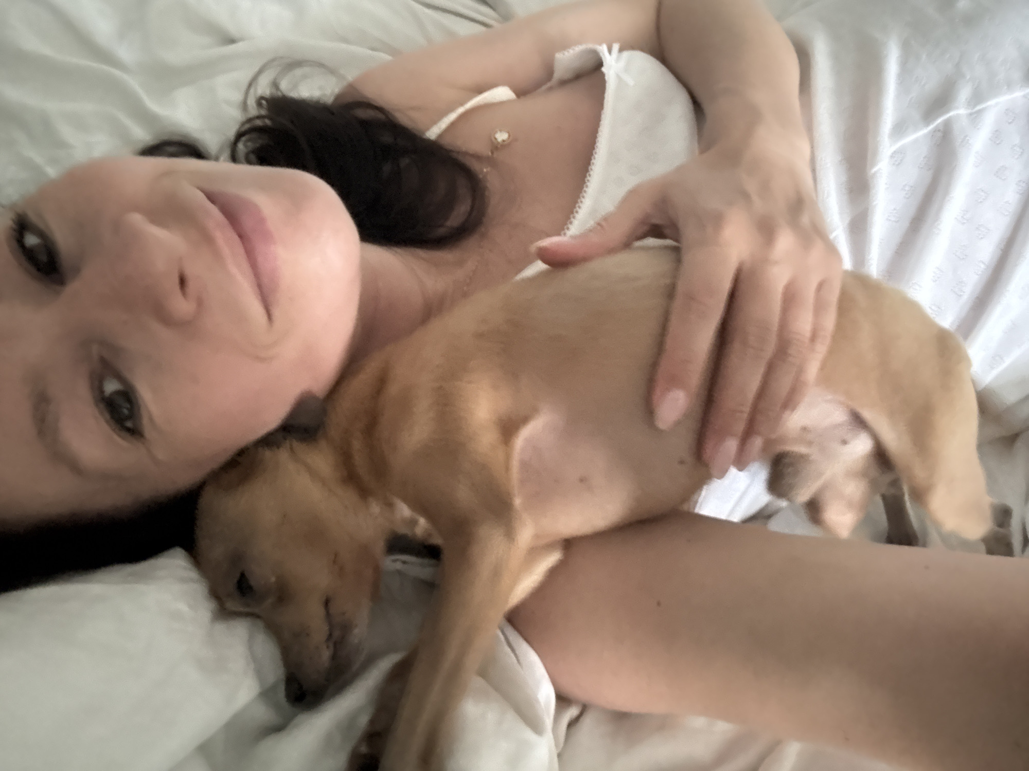 A woman with dark hair and fair skin is lying in a bed covered with white sheets. She is wearing a white top with lace trim and a delicate necklace. She is holding a small, light-brown puppy against her chest. The puppy is lying on its side, appearing to be asleep. The woman is looking at the camera with a slight smile. The location of this image is unknown and unrelated to the city of Chaikovsky, Russia.