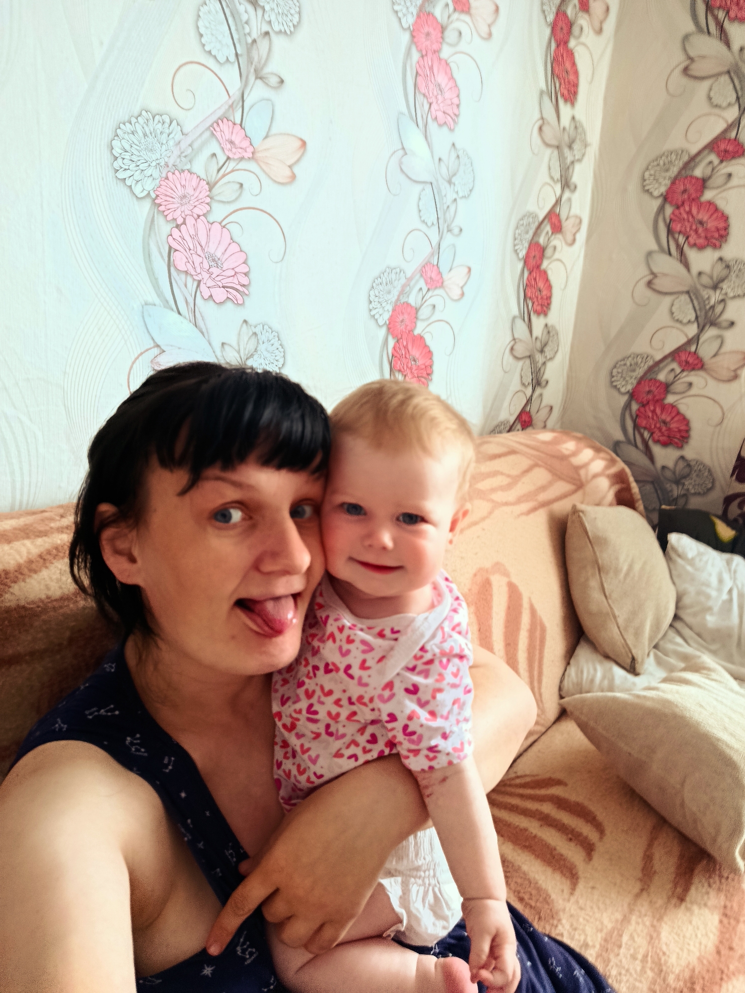A photograph taken indoors shows a woman and a baby. The woman has short, dark hair with bangs, fair skin, and blue eyes. She is sticking her tongue out and looking at the camera. She is wearing a dark blue sleeveless top with white star patterns. The baby is fair-skinned with light blonde hair. The baby wears a white and pink heart-patterned onesie. The woman is holding the baby close. They are sitting on a brown and beige patterned couch, and there are several beige throw pillows behind them. The wall behind the couch is decorated with floral wallpaper in shades of pink, white, and green. The location is Unknown, Unknown.