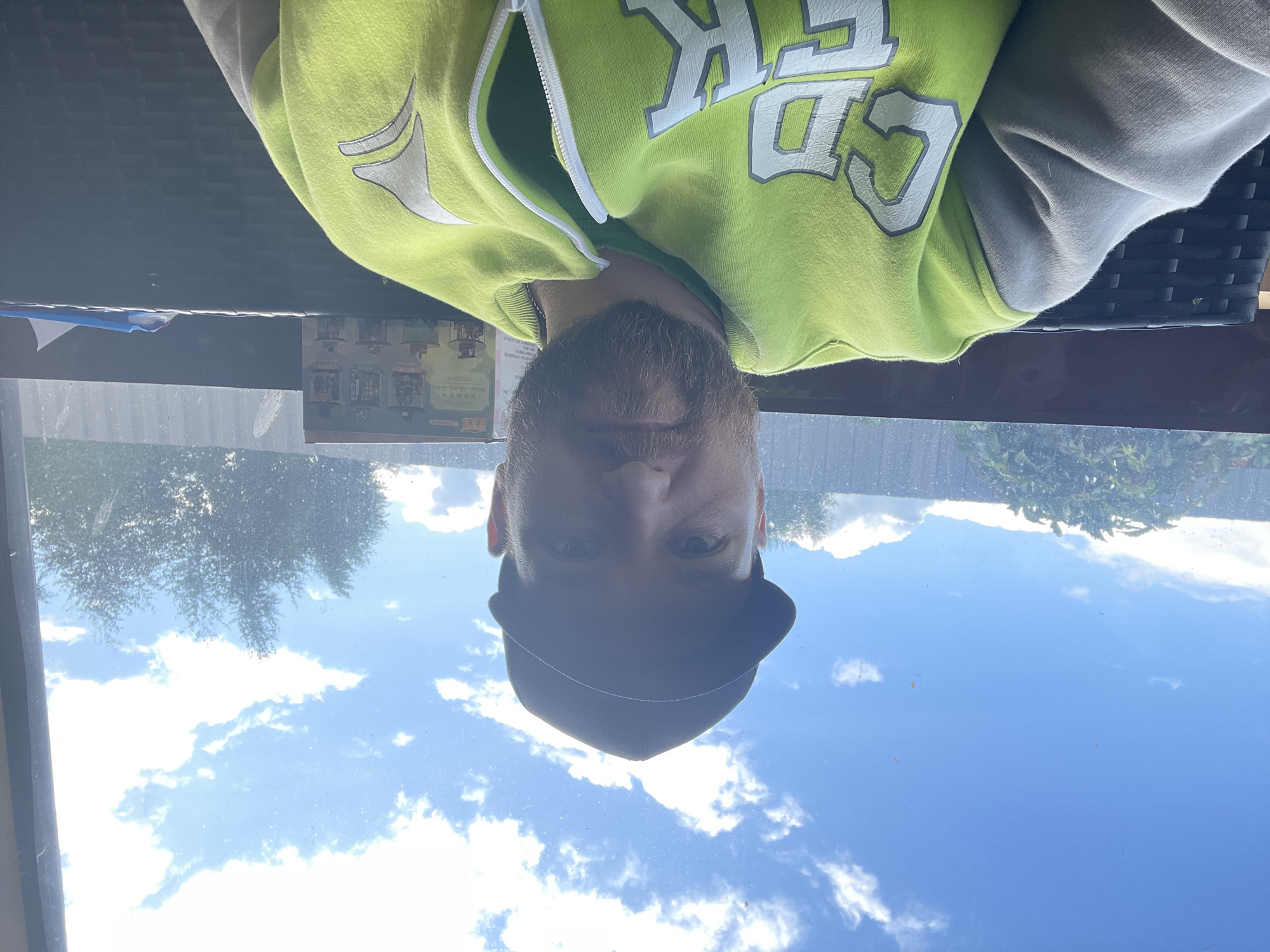 A man is sitting outdoors in front of a window with a bright blue sky and clouds visible. He is wearing a black baseball cap, a lime green hooded sweatshirt with grey sleeves and white lettering on the front, and a green t-shirt underneath. He has a beard and is looking at the camera. In the background, behind him and on a shelf, is a box with images of miniature houses or buildings. The overall scene suggests a casual setting, possibly a backyard or patio, in the city of Kuz'mishchevo, Russia.
