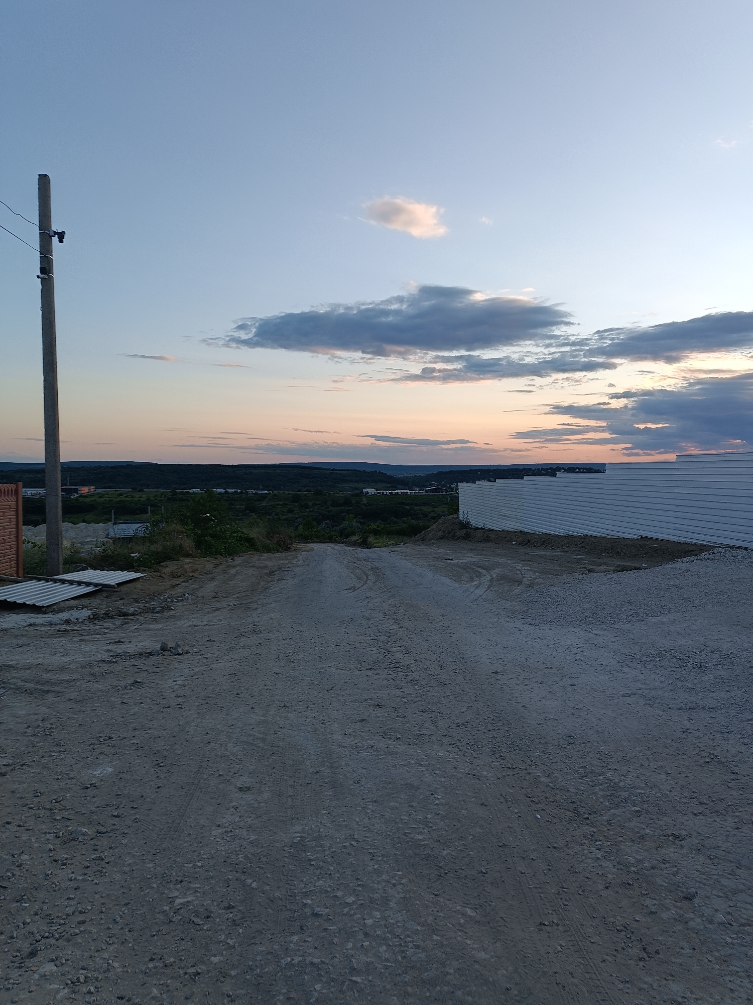 The image depicts an outdoor scene at dusk, looking down a dirt road in Durlești, Moldova. The road is composed of gravel and soil, with visible tire tracks. On the left side of the road, a wooden fence post stands tall, with electrical wires extending from it. Adjacent to the fence post are sheets of corrugated metal. Further down the road on the left, a construction site is partially visible, with some scattered building materials and greenery. On the right side of the road, a long, white corrugated metal fence stretches into the distance. The background reveals a rolling landscape of trees and hills, with a faint horizontal line of buildings or structures in the distance. The sky above is a gradient of pale blue to a soft orange and pink near the horizon, with scattered clouds, indicating the sun is setting. No people are visible in the image.