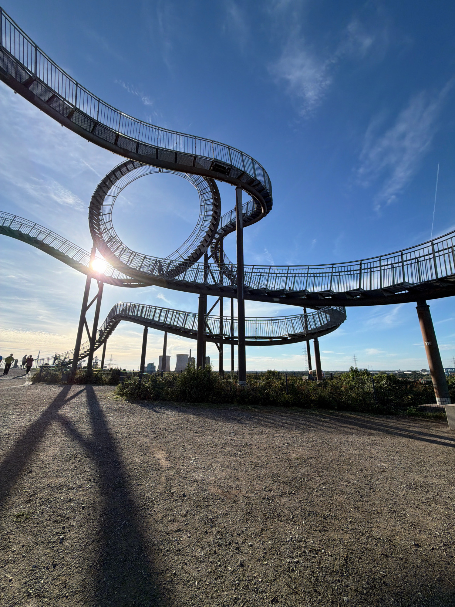 The image depicts the "Tiger and Turtle – Magic Mountain" sculpture, an art installation located on a former slag heap in the Brookhainer Straße district of Duisburg, Germany. The sculpture, made of steel, resembles a roller coaster and allows visitors to walk up and around it. The sun is shining brightly, casting long shadows on the gravel path in the foreground. Several people are visible walking on the sculpture and on the path. In the background, industrial structures, possibly cooling towers of a power plant, are visible against a clear blue sky with scattered clouds. The overall scene is one of public art integrated into an industrial landscape, offering panoramic views.