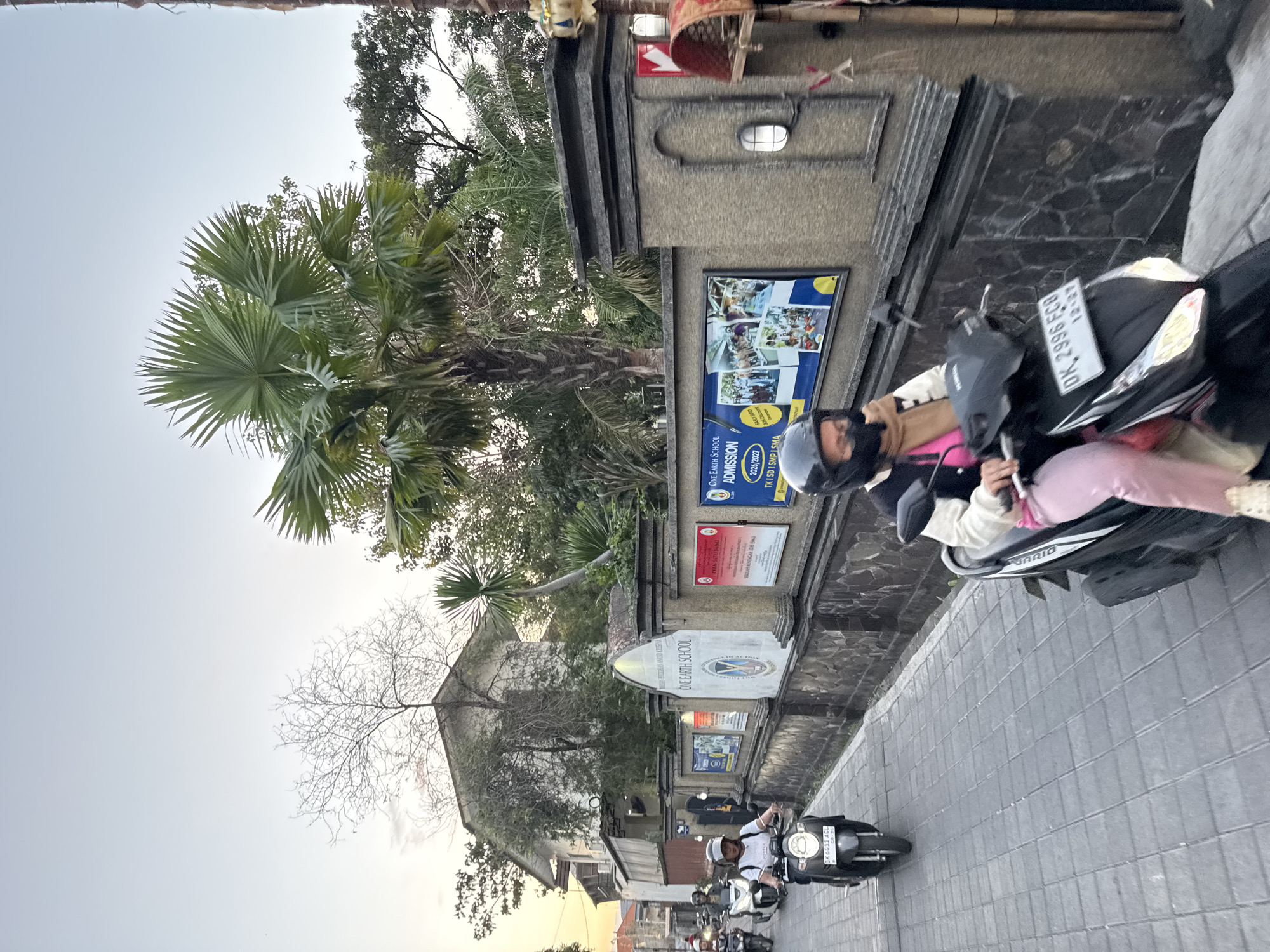 The image shows a street scene in Denpasar, Indonesia, with several people riding motorcycles. The street is paved with gray tiles and is lined with a stone wall. On the wall, there are several signs, including one for "ONE EARTH SCHOOL" with "ADMISSION" and "2024/2027" visible. There are also pictures of students engaged in activities. A woman wearing a helmet and a brown scarf is riding a black motorcycle in the foreground. In the background, another woman on a scooter is visible, along with other motorcycles. A large palm tree is prominent in the upper right portion of the image, and a building with a white facade is visible behind the wall. The sky is overcast and appears to be sunset.