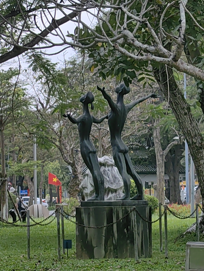 This image depicts a bronze sculpture of two stylized figures positioned outdoors in a park setting within Thuận Hóa Ward, Vietnam. The figures are slender, with elongated forms, raised heads looking upwards, and outstretched arms, standing on a textured, rock-like base that rests upon a dark, rectangular plinth. The sculpture is encircled by a low, dark chain fence with vertical posts, set on a bed of green grass. In the background, beyond the sculpture, an urban environment is visible. Numerous trees with varying foliage and bare branches frame the scene, creating a natural backdrop. To the left, a vibrant red flag of Vietnam, featuring a yellow star, is prominently displayed on a pole near a street. Along the street, vehicles and what appear to be streetlights and additional red banners are discernible. The sky above is overcast, casting a soft, diffuse light over the scene. No active human subjects are clearly identifiable in direct interaction with the sculpture or immediate foreground, though distant figures may be present on the street.