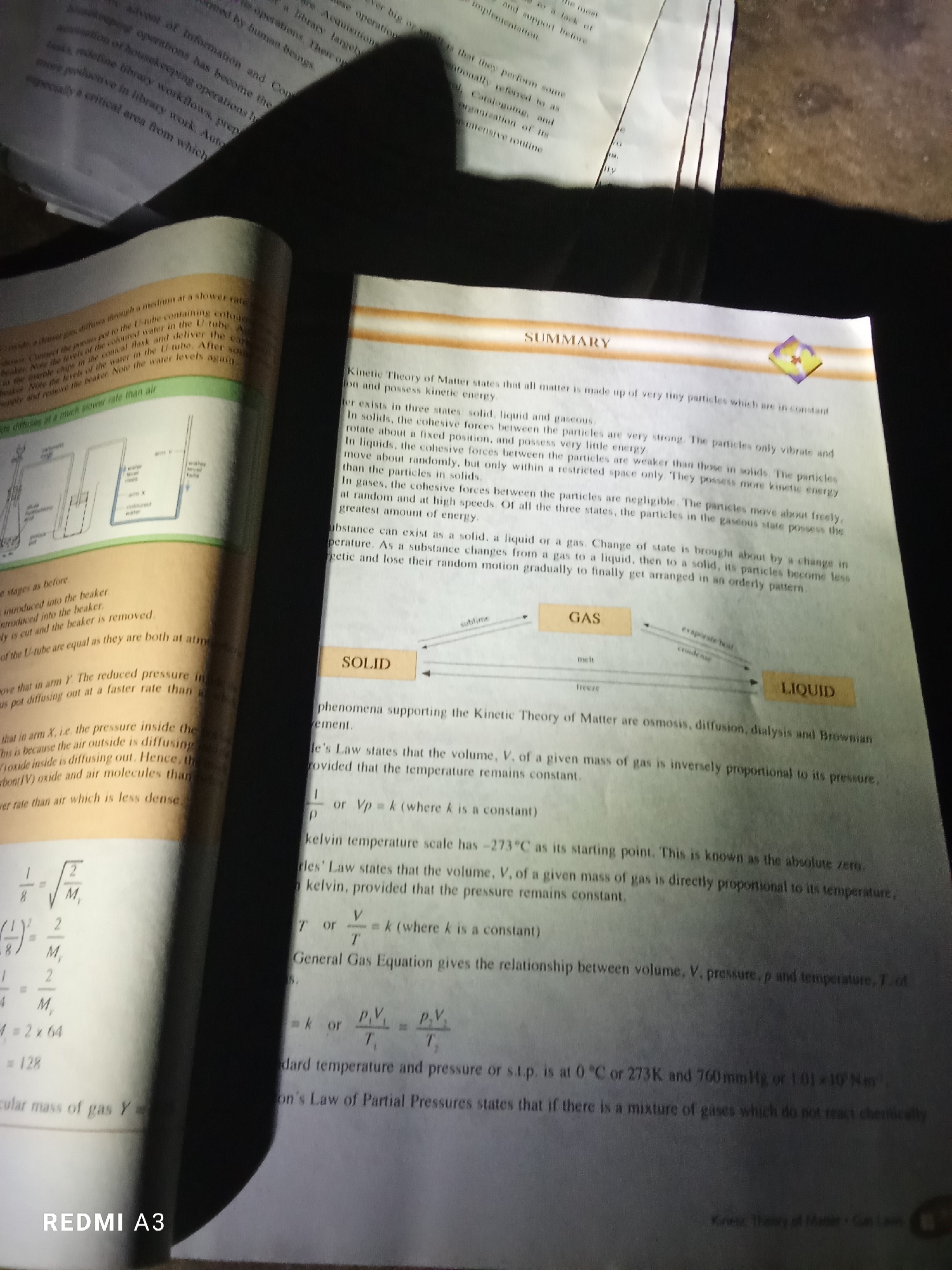 The image shows an open book lying on a surface, with a shadow cast over the right side of the frame. The book appears to be a science textbook, with diagrams and text related to physics or chemistry. The left page of the book displays a diagram illustrating a U-tube experiment, with labels indicating "water level" and "air". There are also some mathematical equations and calculations. The right page is titled "SUMMARY" and discusses the Kinetic Theory of Matter. It explains the properties of solids, liquids, and gases, and includes a diagram showing the transitions between these states (solid, liquid, gas). There is also text detailing various gas laws, such as Boyle's Law and Charles's Law, and their corresponding equations. Visible text includes "SUMMARY", "SOLID", "GAS", "LIQUID", "REDMI A3", and references to scientific terms like "osmosis", "diffusion", and "Brownian movement". The lighting suggests an indoor setting, possibly during the day with artificial light or natural light from a window. No people are visible in the image. The overall impression is of a study session or a page from a textbook being examined.