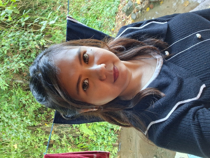 A brunette woman with light skin gazes at the camera with brown eyes and a faint smile. She is wearing a dark, knitted sweater with white trim and pearl-like buttons. Lush green foliage and some bare branches surround her. The background also reveals what appears to be a concrete or stone surface, suggesting an outdoor setting. Part of a dark object with a reddish trim is visible near the bottom left corner.