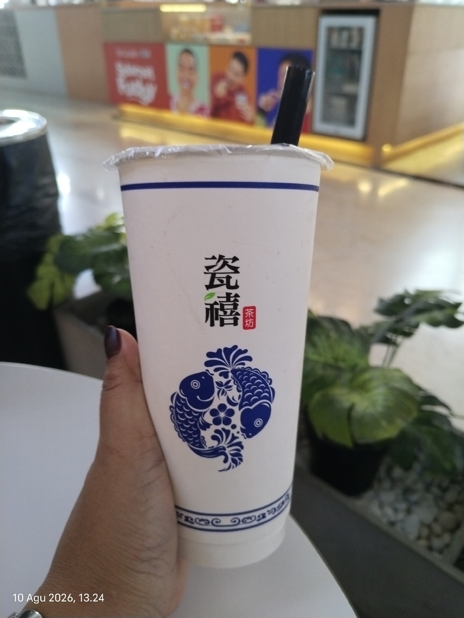A close-up, eye-level shot shows a person's hand holding a white disposable cup with blue accents. The cup has Chinese characters and a blue fish design printed on it, along with a black straw. The background is blurred, revealing a store or cafe interior with a refrigerator and posters. The timestamp "10 Agu 2026, 13.24" is visible in the bottom left corner. This image likely depicts a person enjoying a drink at a cafe or bubble tea shop in East Jakarta, Indonesia.