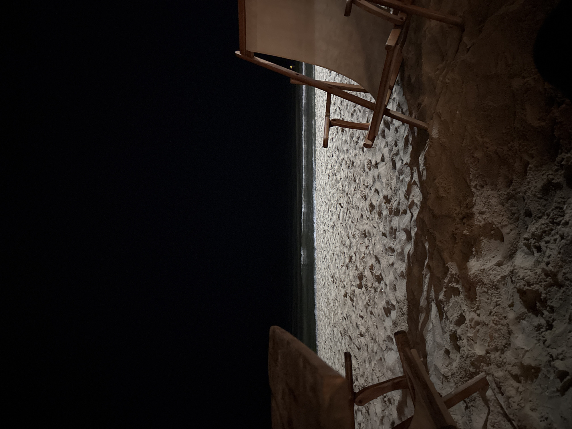 This image captures a nighttime scene on a sandy beach in Varna, Bulgaria. Two wooden beach chairs are positioned in the foreground, facing towards the ocean. The chair on the left is partially visible, with only its upper-left wooden frame and a portion of its surface appearing in the frame. The chair on the right is more fully visible, displaying a wooden frame and a light-colored canvas or fabric seat and backrest, positioned at an angle. Both chairs cast distinct shadows onto the sand, indicating a light source, not visible in the frame, illuminating the beach from a low angle.
The midground consists of a wide expanse of light-colored sand, showing textures and subtle undulations, stretching towards the water's edge. In the background, the ocean is visible with gentle waves breaking onto the shore, distinguishable by faint white foam lines illuminated against the dark water. The sky above the ocean and beach is uniformly black, suggesting a moonless or heavily clouded night with no visible celestial bodies. A small, faint point of artificial light is discernible on the horizon line, slightly to the right of the center, possibly from a distant coastal structure or vessel. No individuals are present in the scene.