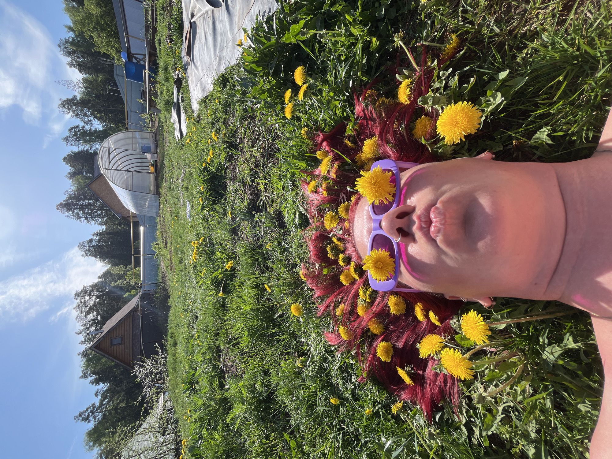 A woman is lying on her back in a grassy field, with dandelions scattered around her hair and on her purple sunglasses. She has red hair and is puckering her lips. Behind her are trees, a building with a greenhouse attached, and a partially visible structure covered in white material. The location is the city of Chaikovsky, Russia.