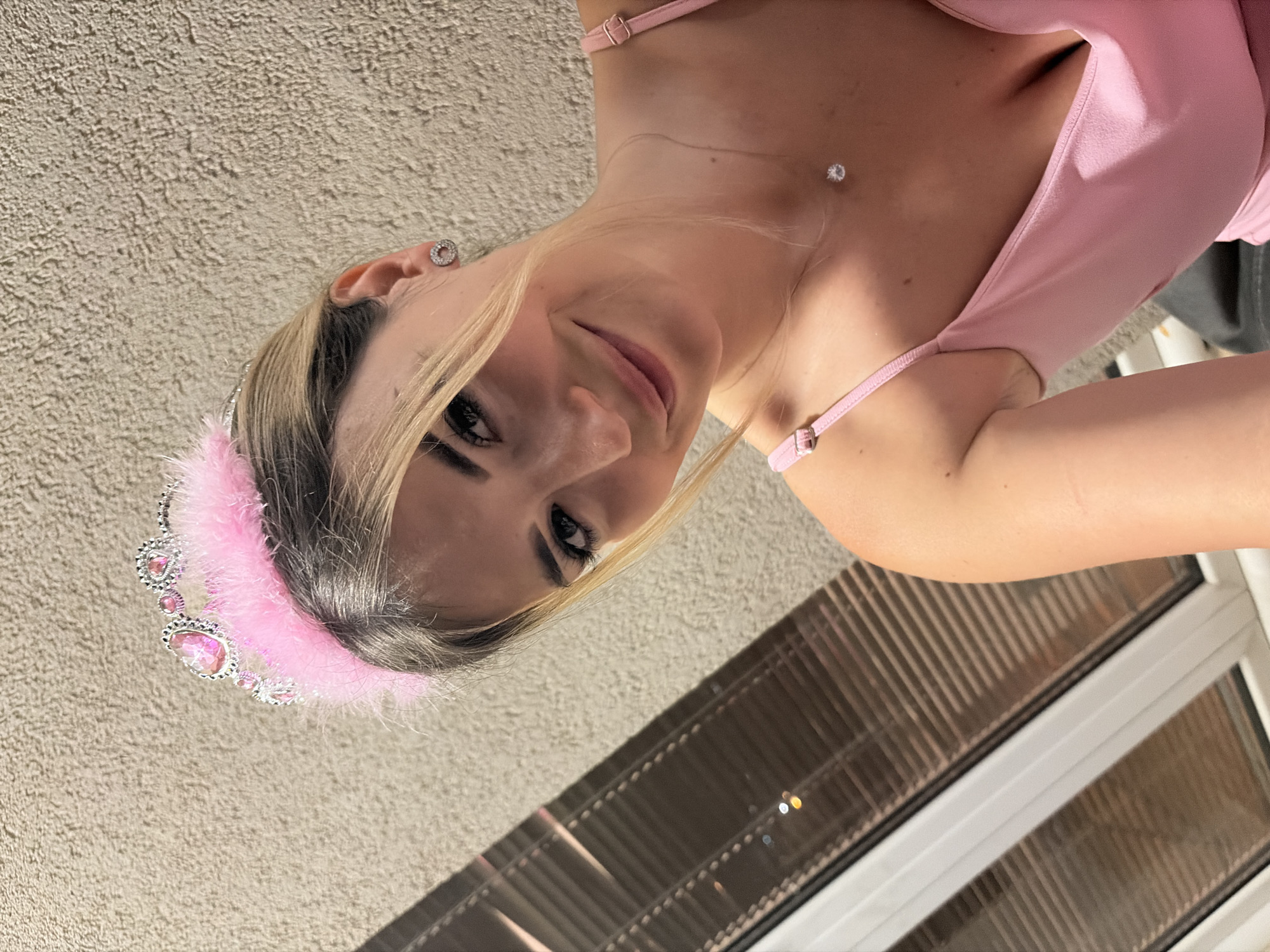 A blonde woman with a pink fluffy tiara and a pink dress smiles at the camera. She is wearing a diamond necklace and stud earrings. The background is a beige wall and a window with blinds. The image appears to be taken indoors, possibly in an apartment or hotel room, in Budva, Montenegro, based on the provided context.