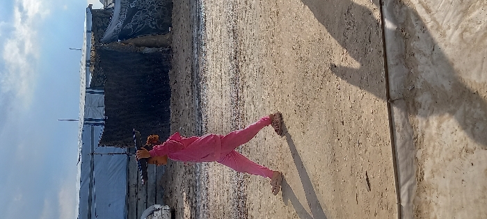 A young girl walks left to right across a sunlit, uneven dirt path in Khan Yunis, Palestinian Territory. She wears a pink tracksuit and light-colored sandals, carrying a dark, rectangular plastic object, possibly a tray or lid, on her head, stabilized by her left hand. Her hair is dark and curly. Behind her, makeshift structures are visible: to the left, a shelter constructed from white or silver tarp-like material over a frame; to the right, a darker, rustic shelter made of coarse, textured materials. A portion of a white vehicle is partially visible on the far left. The sky is partly cloudy blue, casting distinct shadows on the ground.