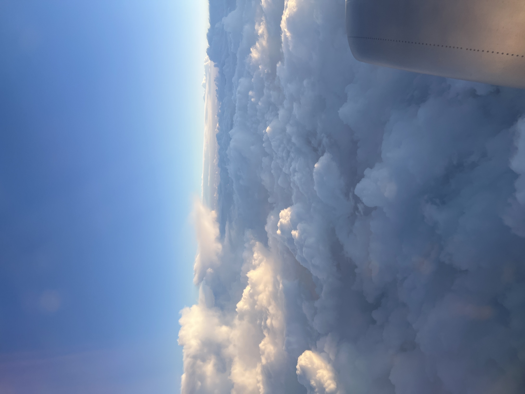 The image is a view from an airplane window. The scene shows a clear blue sky on the left and a vast expanse of fluffy, white clouds on the right. The top of an airplane wing is visible in the upper right corner. The lighting suggests it is daytime. The location of Erkheim, Germany is not explicitly evident within the scene.