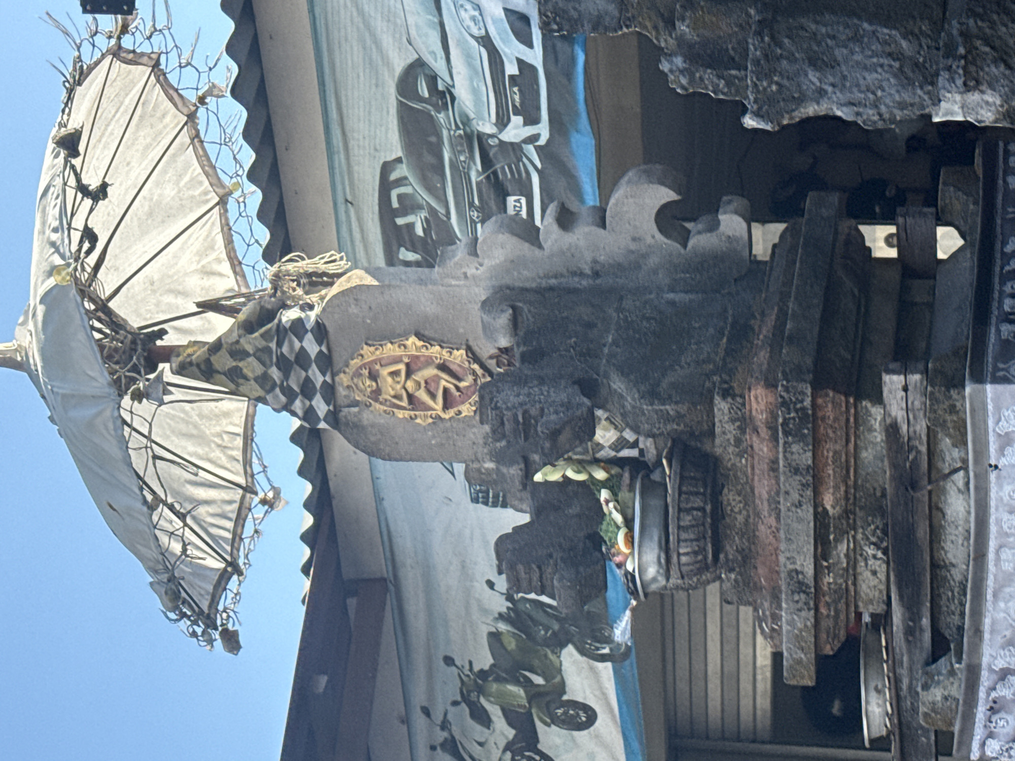 The image shows a stone altar with a traditional Balinese umbrella, adorned with a checkered cloth. In the background, a banner depicts motorcycles and cars. The altar is decorated with offerings, including fruits and flowers. The scene appears to be in Denpasar, Indonesia, likely in front of a shop or business, given the banner. The sky is clear and blue, indicating a sunny day.