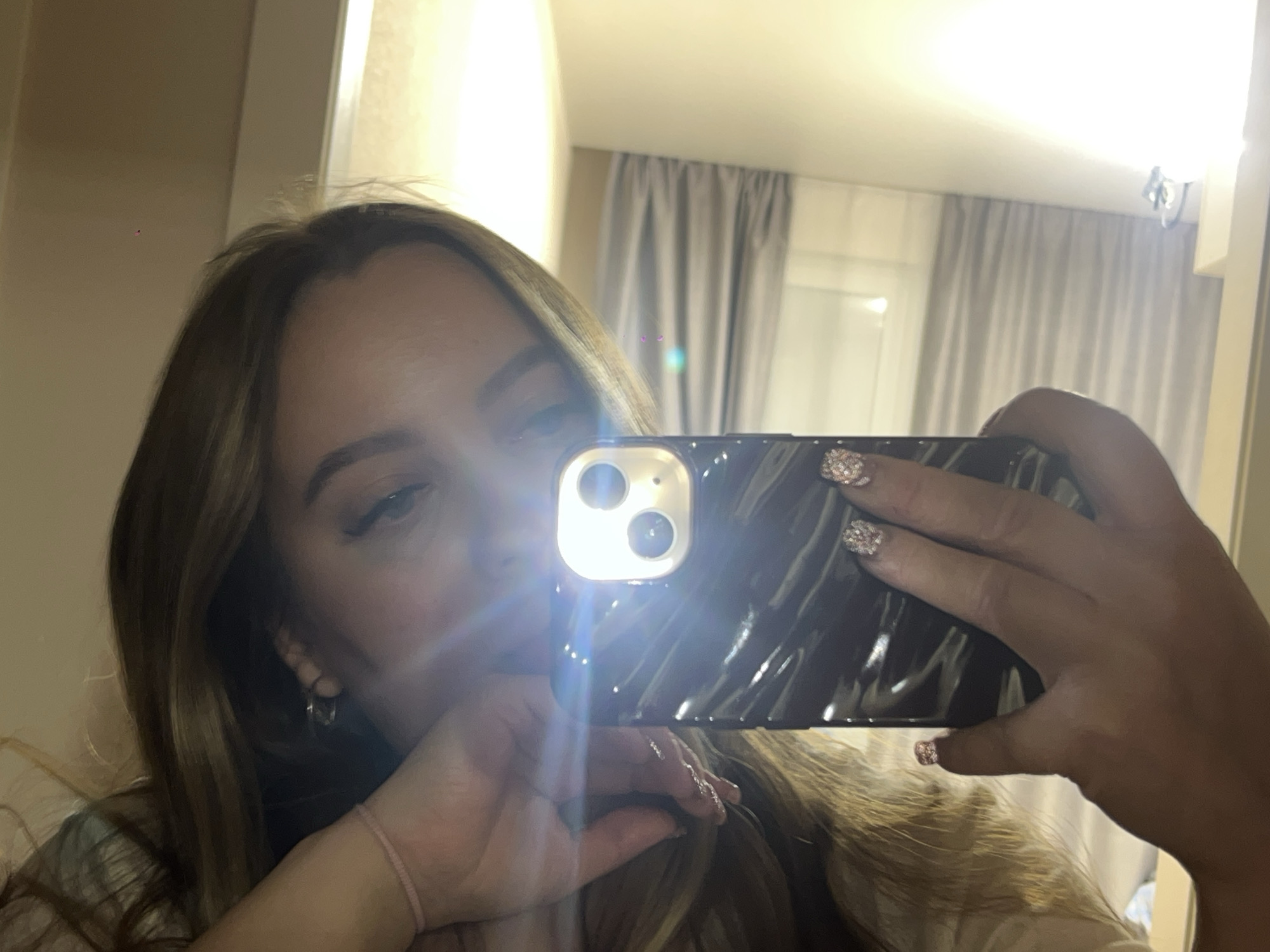 A young woman is taking a selfie in a room with beige walls and light gray curtains. She is holding a black smartphone with a decorative, textured case. The phone's flash is on, creating glare. She has long brown hair with blonde highlights. Her nails are decorated with glitter. She is wearing a thin bracelet on her left wrist and a hoop earring in her left ear. The reflection in the mirror shows a room interior, including a wall lamp. There is no information to place the location in Krasnodar, Russia.