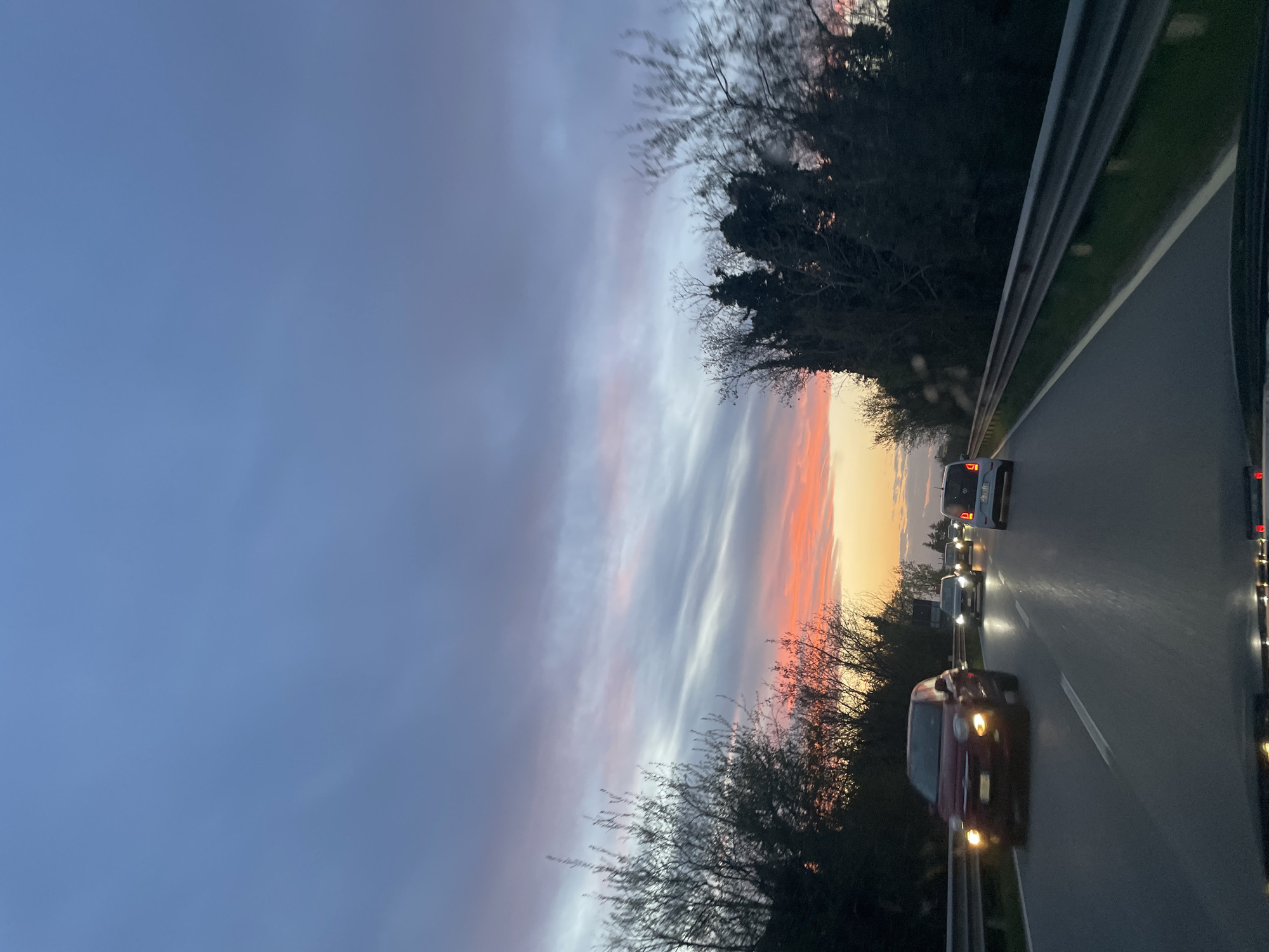 The image captures a view from a moving vehicle, likely on a highway, in San Giovanni al Natisone, Italy, at dusk. The sky displays a gradient of colors, from a deep blue at the top to hues of pink, orange, and white near the horizon. Trees line the edges of the road. Several vehicles with headlights on are visible on the road ahead, suggesting moderate traffic. A guardrail runs alongside the road on the right side of the image.