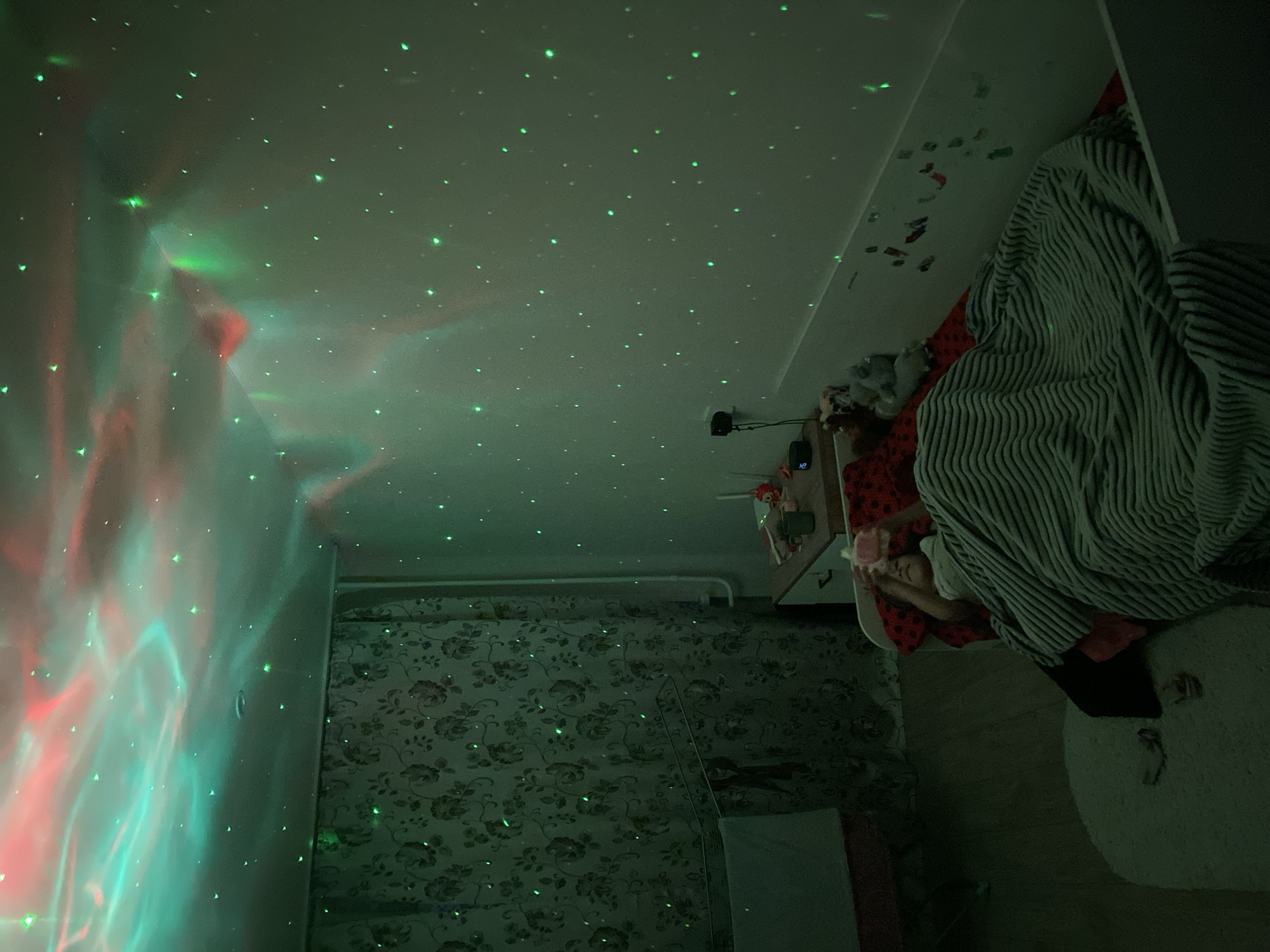 A dimly lit bedroom in Chaikovsky, Russia. The ceiling is covered in green star-shaped lights and swirling aurora-like projections in red and green. Wallpaper with a dark floral pattern covers one wall. A child lies in bed under a striped blanket. A white, round rug is on the floor. A small bedside table is adjacent to the bed.