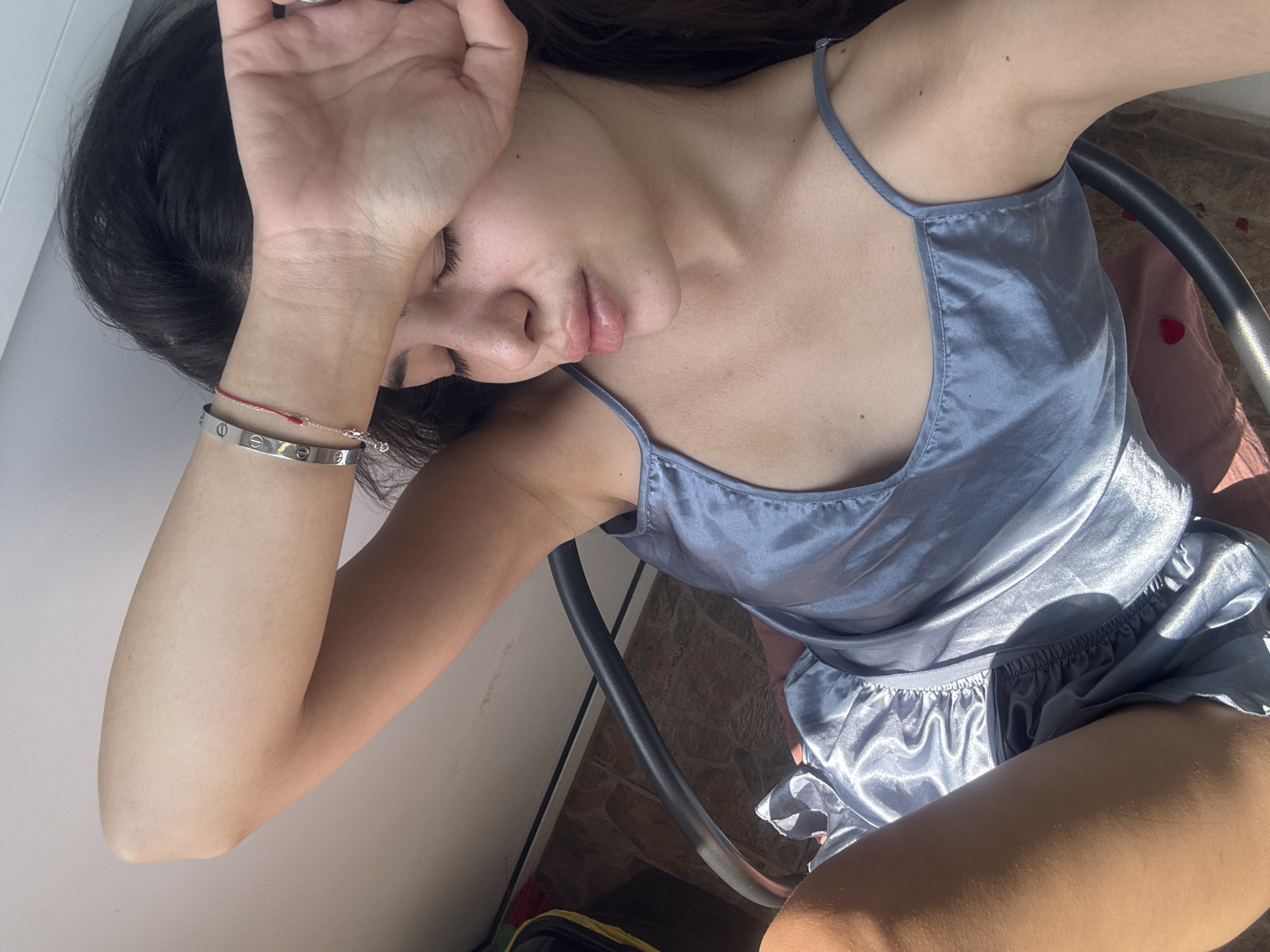 A young woman is depicted in a moment of repose, likely indoors or on a balcony, in Plovdiv, Bulgaria. She is lying down, seen from an overhead perspective, with her right arm raised, partially covering her face as if shielding her eyes from the bright sunlight or simply resting. Her eyes are closed, and her lips are slightly parted, conveying a sense of drowsiness or peaceful slumber.
She is wearing a light blue or silver-grey satin-like camisole top and matching shorts, suggesting loungewear or pajamas. On her right wrist, she accessorizes with a silver-toned bracelet featuring screw-like markings, resembling a Cartier Love bracelet, along with a delicate red string bracelet.
The scene is bathed in strong, direct sunlight, indicating daytime. A dark, possibly metal, chair frame is visible, and the background reveals a patterned floor, likely stone or tiles, with some scattered red petals (possibly rose petals) resting on a pink fabric or cushion. The overall atmosphere is serene and relaxed, emphasizing a moment of quiet comfort. No explicit text is clearly legible beyond the design details on the bracelet.