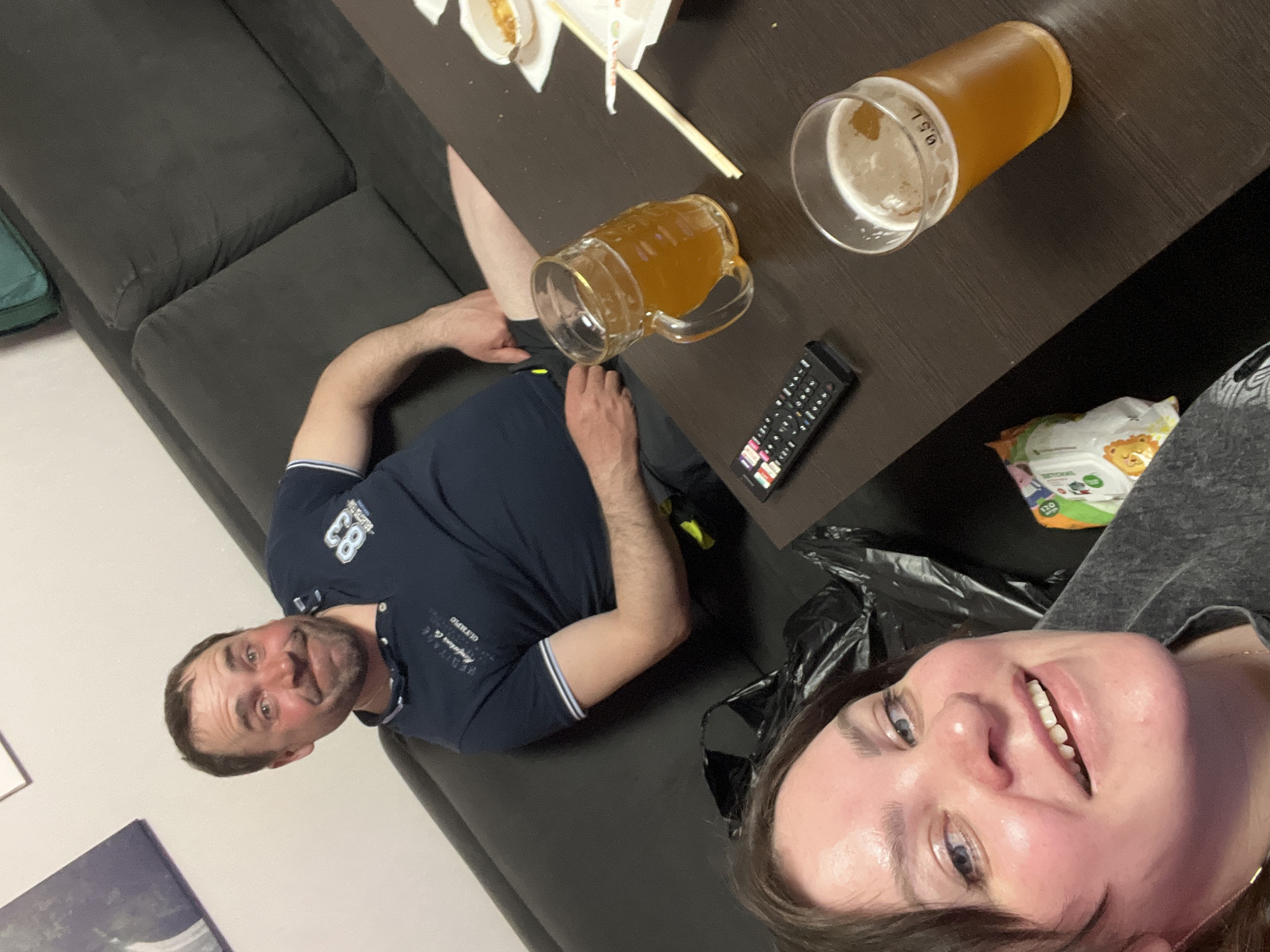 The image captures a casual indoor scene, likely taken with a wide-angle lens or a selfie perspective. A man in a navy blue t-shirt with white accents is seated on a dark gray sofa. He is looking directly at the camera with a slight smile. Beside him, a woman is positioned closer to the camera, also smiling.
In front of them is a dark brown coffee table. On the table, there is food in a white container, a set of chopsticks, a 0.5-liter glass of beer, a beer mug filled with beer, and a black remote control. There is also a bag of chips. The background includes the dark gray sofa and hints of a wall with an abstract painting.
While the image does not confirm the location in the city of Perm, Russia, that could be the location.