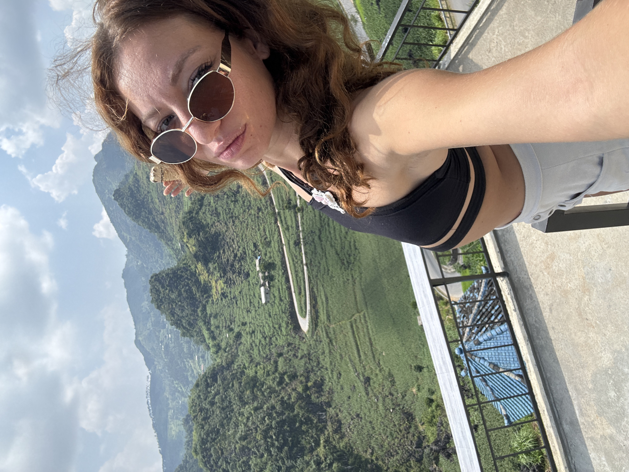 A woman with brown hair and sunglasses takes a selfie in Lũng Phìn Commune, Vietnam. She is wearing a black top and light-colored shorts. Behind her are green, mountainous hills and a sky with light clouds. A curved road is visible in the green landscape. A railing is also visible next to her.