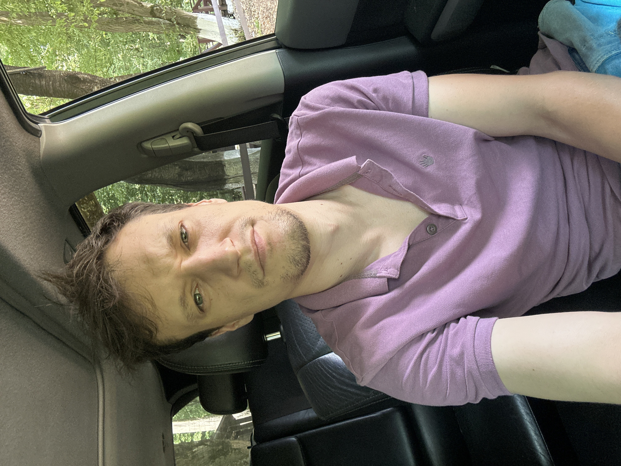 A man is seated in the driver's seat of a car, his head and upper torso visible in the foreground. He is wearing a light purple polo shirt. The interior of the car is dark, with leather seats visible in the background. Through the windows, a lush green forest can be seen, suggesting a rural or natural setting. The man's expression is neutral, and he appears to be looking towards the camera. The overall lighting indicates it is daytime.