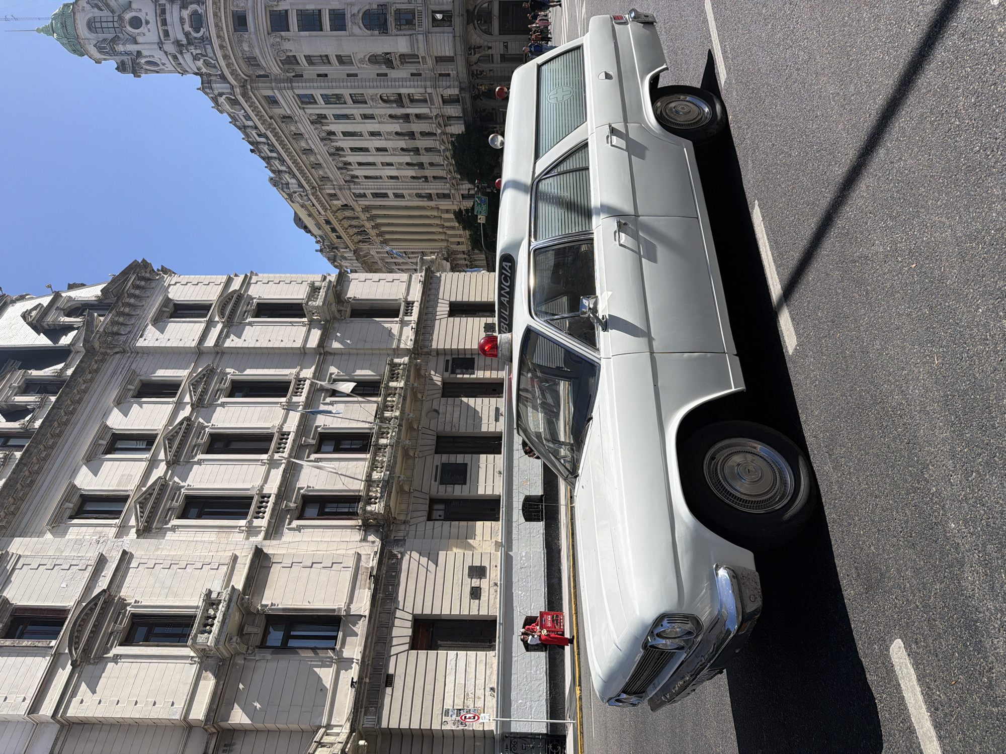 A white ambulance with the word "AMBULANCIA" written on its side is parked on a street in Buenos Aires, Argentina. Two red garbage bins and two people in red and white uniforms stand behind the ambulance. The street surface is asphalt. Imposing historical buildings line the street. One is a lighter-colored building with many windows and intricate architectural details. Another grand building with a domed structure is visible in the background. The sky is clear and blue.