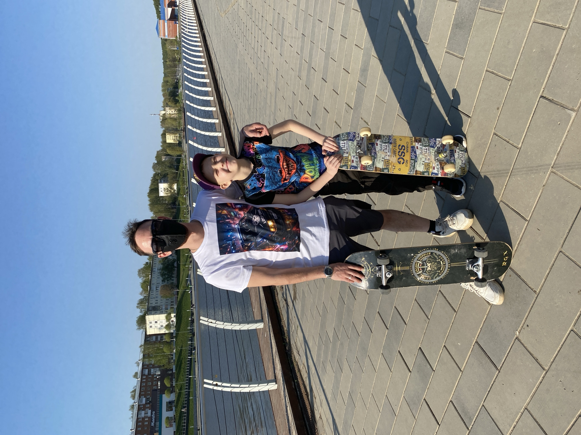 On a sunny day in Votkinsk, Russia, an adult male wearing a white t-shirt with a graphic print, black shorts, white sneakers, and a black face mask stands beside a younger male, presumably a child, who is lying on his back, smiling and wearing a purple baseball cap and a colorful patterned t-shirt. Both individuals are positioned on a grey-paved surface, each with a skateboard beside them. The older male holds his skateboard in his hand, while the younger male is resting on his.
In the background, there is a structure with white rails and a dark surface, likely a walkway or bridge, overlooking buildings and greenery. The sky is a clear blue. Based on the OCR text, there are various details on the skateboards, including references to "RGX", "ASGARD", "SSC", "NORTH CARO", and the words "LOOP ONE". The lighting suggests it is midday.
