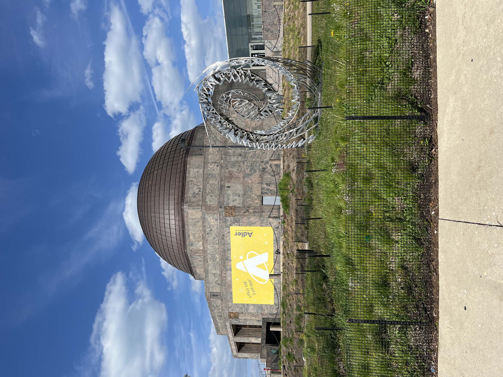 The image shows the Adler Planetarium in Chicago, Illinois, USA. The building has a distinctive dome-shaped roof and is constructed of stone. In front of the building is a metal sculpture. There is a yellow sign with the planetarium's logo and the words "Space for Everyone!" A black wire fence is visible near a concrete walkway. The sky is blue with scattered white clouds.