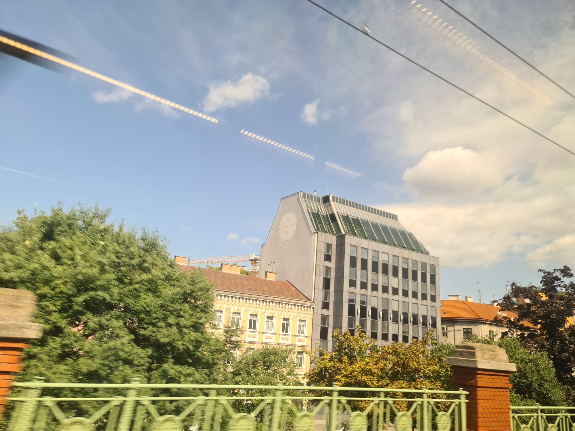 The image depicts a city scene, likely from a moving vehicle, showing a modern office building with a glass facade and a pitched, glass-covered roof. Adjacent to it is a yellow, older building with classical architectural elements. Green trees are abundant in the foreground and mid-ground, and a bright orange brick structure with a decorative green railing is visible. In the background, a construction crane is present. The sky is blue with scattered clouds. The image shows a section of the Ringstrasse in Vienna, Austria, near the Stock Exchange building. The time of day appears to be daytime, with sunlight casting shadows. There are no people visible in the image. The reflections of lights and possibly electrical wires are visible on the surface from which the photo was taken, suggesting it was shot through a window.