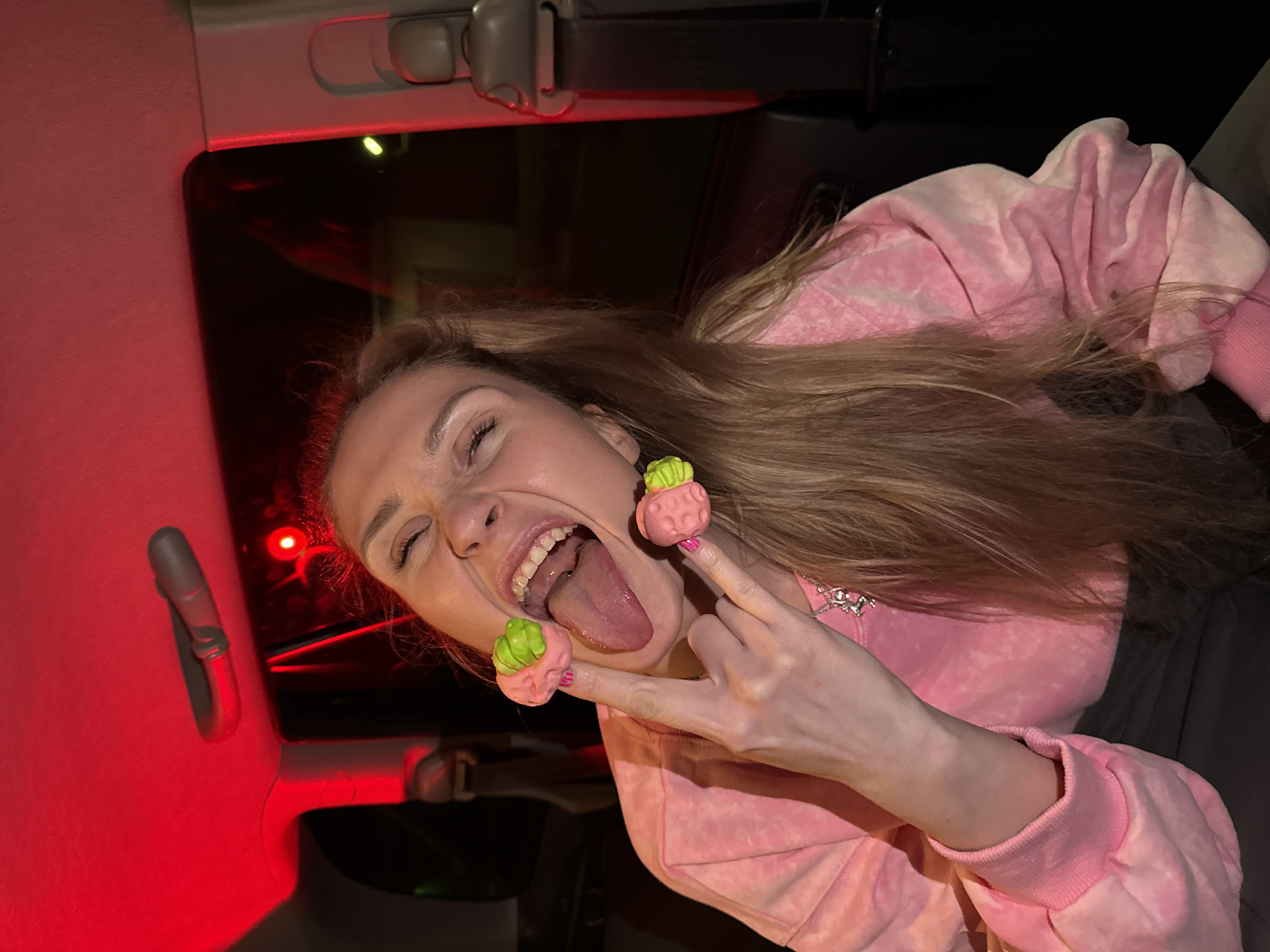 A woman is inside of a car, sticking her tongue out, and holding up her fingers. The car is in Chaikovsky, Russia. The woman is wearing a pink sweatshirt and has long, blonde hair. She has novelty decorations on her fingers that look like miniature cabbages. The interior of the car is visible, including the ceiling, a seatbelt, and the red interior light. The background outside of the car window is dark.