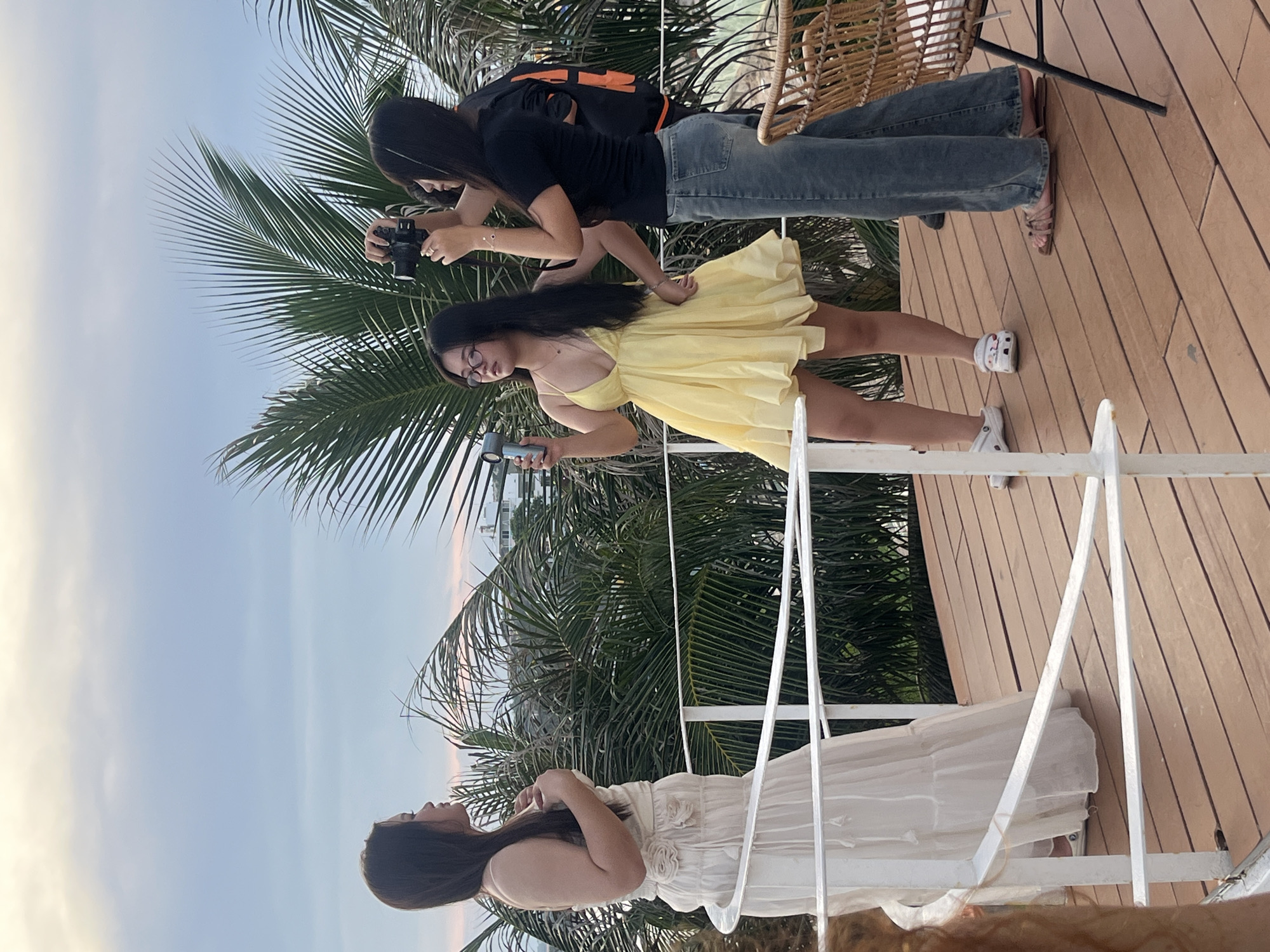 Three young women are on a wooden deck, likely a balcony or patio, with palm trees and a hazy sky in the background. The woman in the foreground is wearing a flowing, cream-colored dress and is looking up and to her left. She has long, dark hair and appears to be engaged in conversation or observing something.
Behind her, another woman is wearing a bright yellow, spaghetti-strap dress. She is holding a camera and seems to be taking a photo of the woman in the cream dress. She has dark hair and is wearing glasses.
The third woman, furthest back, is wearing a black t-shirt, jeans, and a backpack. She is also holding a camera and appears to be taking a photo. She is wearing sandals.
The scene suggests a casual outing, possibly a photoshoot or a group of friends enjoying a scenic location. The lighting suggests it's either early morning or late afternoon, with soft, diffused light. The location cues of palm trees and the general ambiance are consistent with a tropical or subtropical coastal setting like Mũi Né, Vietnam. The mood appears relaxed and happy. No visible text is present in the image.