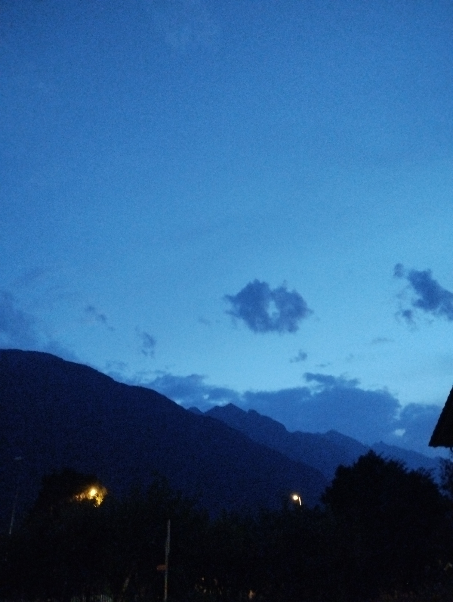 This is a vertical, full-shot, eye-level, low-light photograph of a mountain range at dusk. The sky is a deep, rich blue with wispy clouds scattered across it. The mountains are dark silhouettes against the sky, with several layers visible. In the foreground, there are dark trees and bushes. A single yellow streetlight is visible amongst the trees on the left side of the frame, and another streetlight is visible in the middle ground. The image is grainy, suggesting it was taken in low light conditions. The location is unknown.