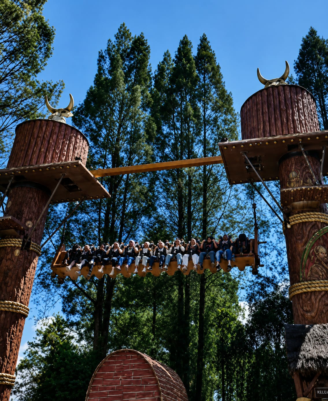 In Berastagi, Indonesia, a group of people are seated on a pendulum ride at an amusement park. The ride features two large, wooden, totem-like structures, each topped with golden bull horns, supporting a horizontal beam from which the pendulum hangs. The people, dressed casually, are positioned on the pendulum, facing forward and appearing to be in mid-swing. The background is dominated by tall green trees and a clear blue sky. A small, red, brick-like structure is visible in the lower portion of the image, partially obscured by foliage. The ride's design has a rustic, possibly Viking-inspired, aesthetic.