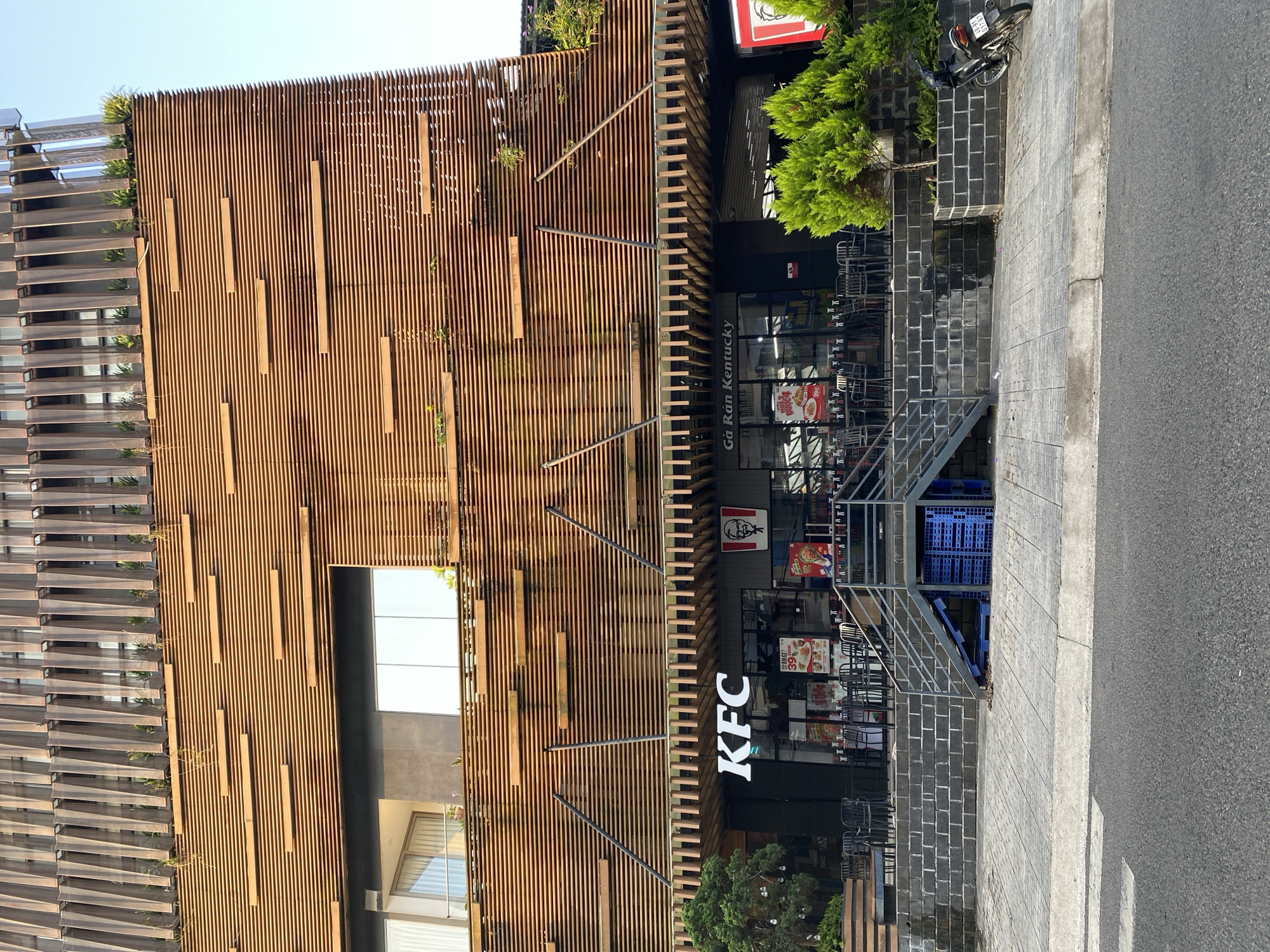 The image depicts a KFC restaurant in Da Lat, Vietnam. The building's facade features a modern design with vertical wooden slats and some greenery. The restaurant entrance is accessible via a staircase. The KFC logo is prominently displayed. A motorcycle is parked on the sidewalk. The street is paved and appears relatively clean.