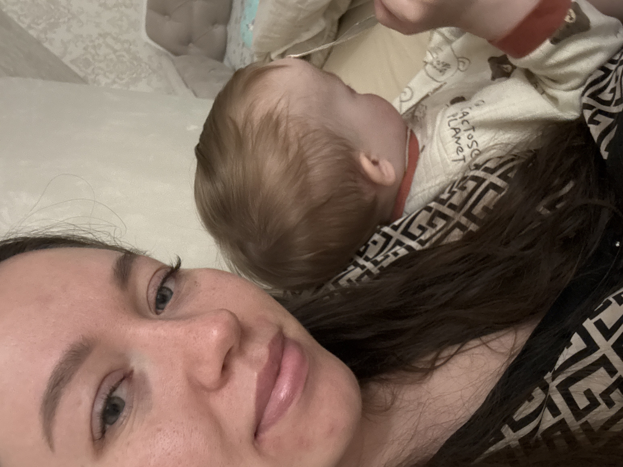 A selfie shows a woman with pale skin and dark hair next to a baby with light brown hair. The baby is lying on its back, partially on the woman's hair. The woman is looking at the camera with a slight smile. The baby is wearing a light-colored outfit with cartoon drawings and red trim. The woman is wearing a black and white patterned top. The background includes a light-colored bedspread and headboard. The scene likely takes place indoors in a private residence. The location, Ферма, Russia, is provided.