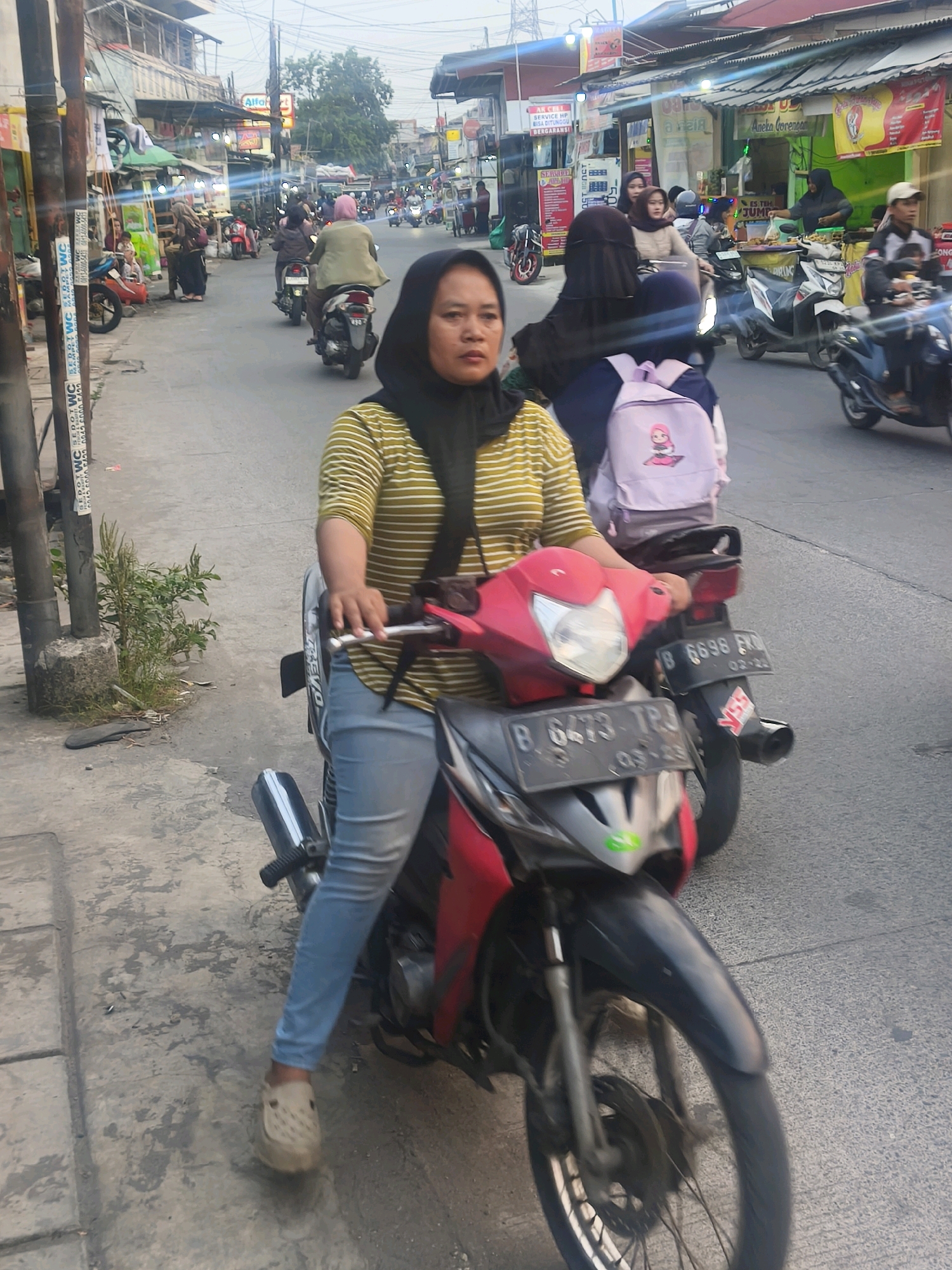 In Bekasi, Indonesia, a woman wearing a black hijab and a striped yellow and white shirt is on a red and black motorcycle. She has on light blue jeans and beige clogs. She is looking forward. The motorcycle is facing the viewer and is on a street with other motorcycles and people. There are shops and buildings on both sides of the street, and the sky is overcast. The license plate on the front of the motorcycle is obscured. The license plate on the back of the motorcycle is visible and reads "B 6696 FRN".