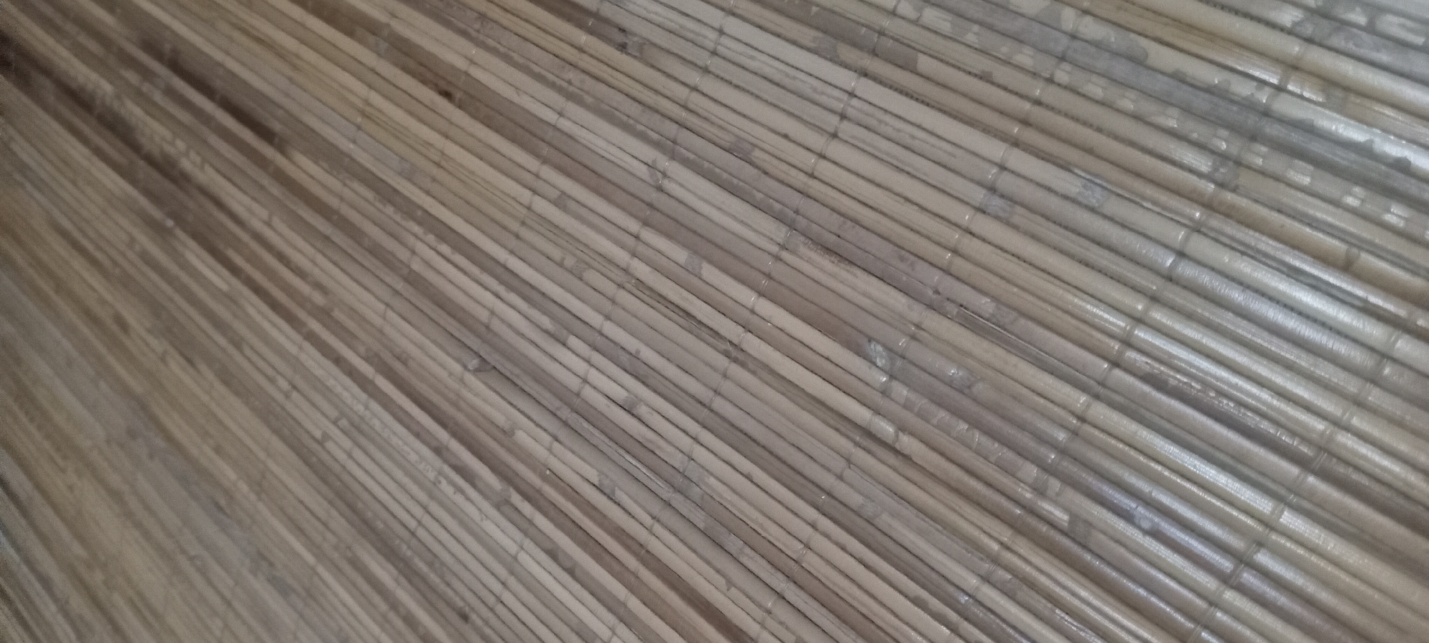 The image shows a close-up, angled view of a woven bamboo mat. The bamboo stalks are thin, light brown to tan in color, and are arranged in parallel lines. The texture appears smooth and somewhat glossy, with visible nodes or joints along the stalks. The lighting creates subtle shadows and highlights, emphasizing the ribbed texture of the material. The perspective suggests the mat is laid out flat, possibly as a floor covering or a part of furniture. There is no indication of a specific location within Sindangwangi, Indonesia, as the image focuses solely on the material itself without any surrounding context.