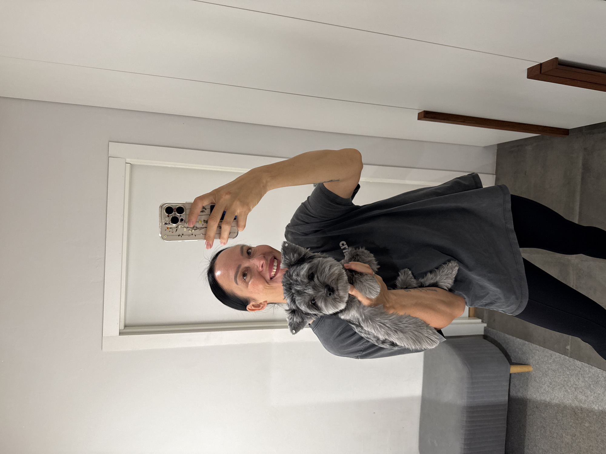 A woman with dark hair pulled back is taking a selfie in a mirror, holding a small gray dog. She is wearing a dark gray t-shirt and black leggings. The dog is held in her left arm, and she is holding her phone in her right hand. The background appears to be an interior space with white walls, a doorway, and a closet. There is also a gray upholstered bench with wooden legs visible to the left. The image does not contain any elements that suggest it is located in the city of Chaikovsky, Russia.