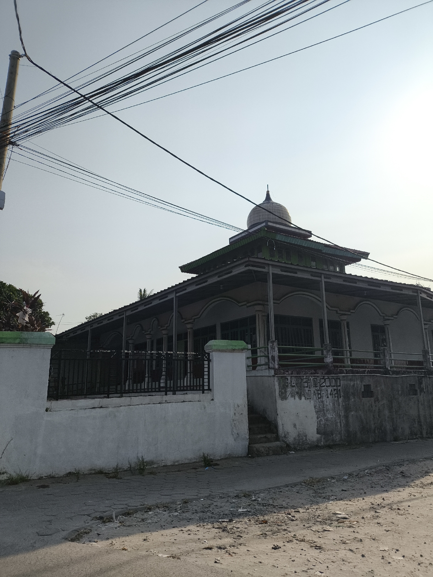 The image shows a mosque with a dome, located in Cilegon, Indonesia. The mosque has a traditional architectural style with a wide, covered veranda supported by pillars. A black metal fence encloses the mosque's property, and a white wall borders the front. The ground in front of the mosque is paved with cracked concrete and scattered debris. Overhead, a tangle of power lines crisscrosses the sky. The overall scene suggests a modest, possibly older, religious building in a typical urban or semi-urban setting in Indonesia. The lighting indicates it is daytime, with the sun casting shadows. There are no people visible in the image.