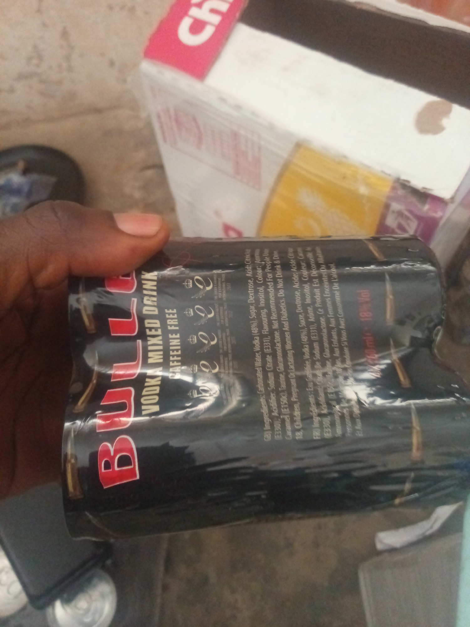 A hand, with dark skin and a short fingernail, holds a multi-pack of four cans of "ULTRA VODKA MIXED DRINK CAFFEINE FREE". The cans are black with red and white lettering and are wrapped in clear plastic. The packaging is slightly creased. In the background, a portion of a white box with pink lettering is visible, along with a yellow cylindrical object, also in plastic wrapping. The scene appears to be outdoors, with a rough, sandy surface visible on the ground. The lighting suggests it is daytime.