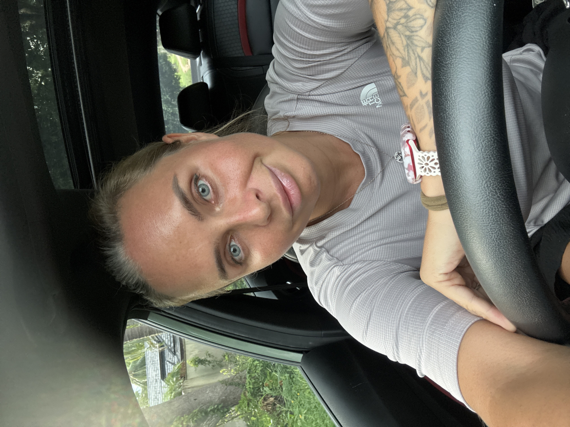 A woman with blonde hair pulled back and piercing blue eyes smiles at the camera from the driver's seat of a car. She is wearing a grey t-shirt with a white logo on the left chest. Her right arm rests on the steering wheel, revealing a large tattoo on her forearm and a decorative bracelet on her wrist. In the background, through the car window, lush green foliage and a roofline are visible, suggesting a tropical environment. The interior of the car shows black upholstery and part of the sunroof. The image appears to be taken during the daytime, with natural light illuminating the scene. The location is identified as Baan Harn, Thailand.