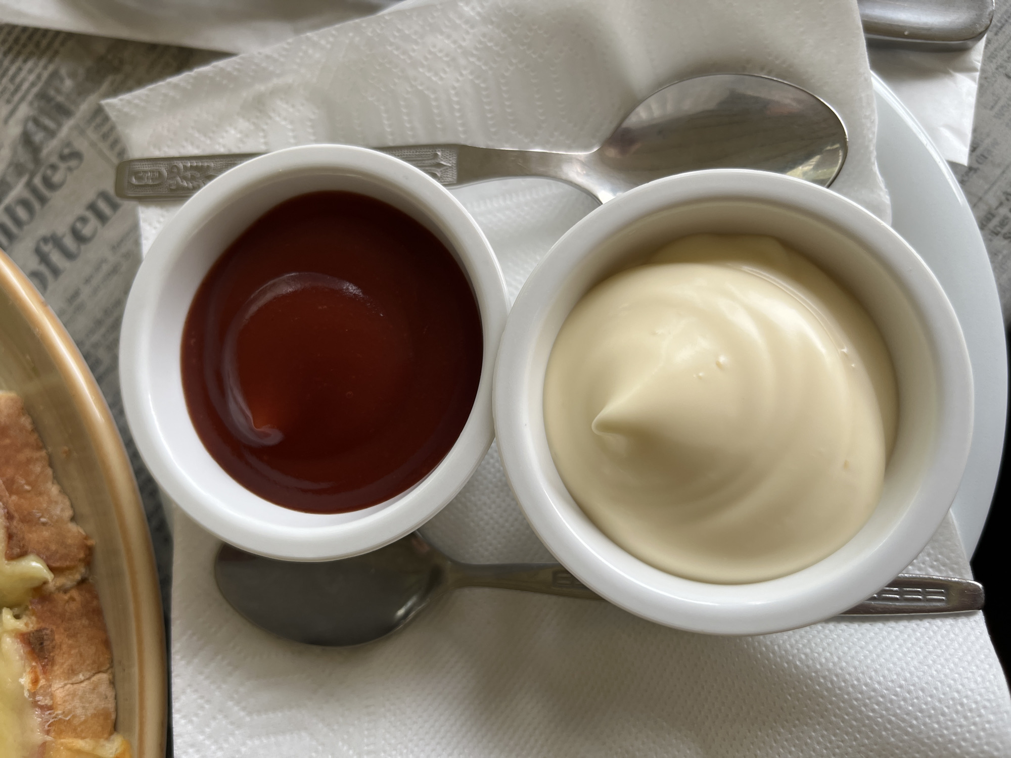 In a close-up, overhead shot, two white ramekins are prominently displayed. The upper ramekin contains a deep red substance, likely ketchup, while the lower ramekin holds a creamy, pale yellow substance, presumably mayonnaise. Both ramekins are positioned on a white, textured napkin. To the left of the ketchup ramekin, a silver spoon rests on the napkin. A second silver spoon is visible to the right of the mayonnaise ramekin, also on the napkin. In the background, a portion of a plate with food, possibly toast with melted cheese, is visible at the top of the image, along with a blurred surface with printed text. The scene appears to be set on a table, with the background suggesting a casual dining or cafe environment. The overall impression is one of food preparation or service, with condiments ready for use. The image does not provide any information about the city of Rudozem, Bulgaria, or any specific details regarding people, actions, or interactions.