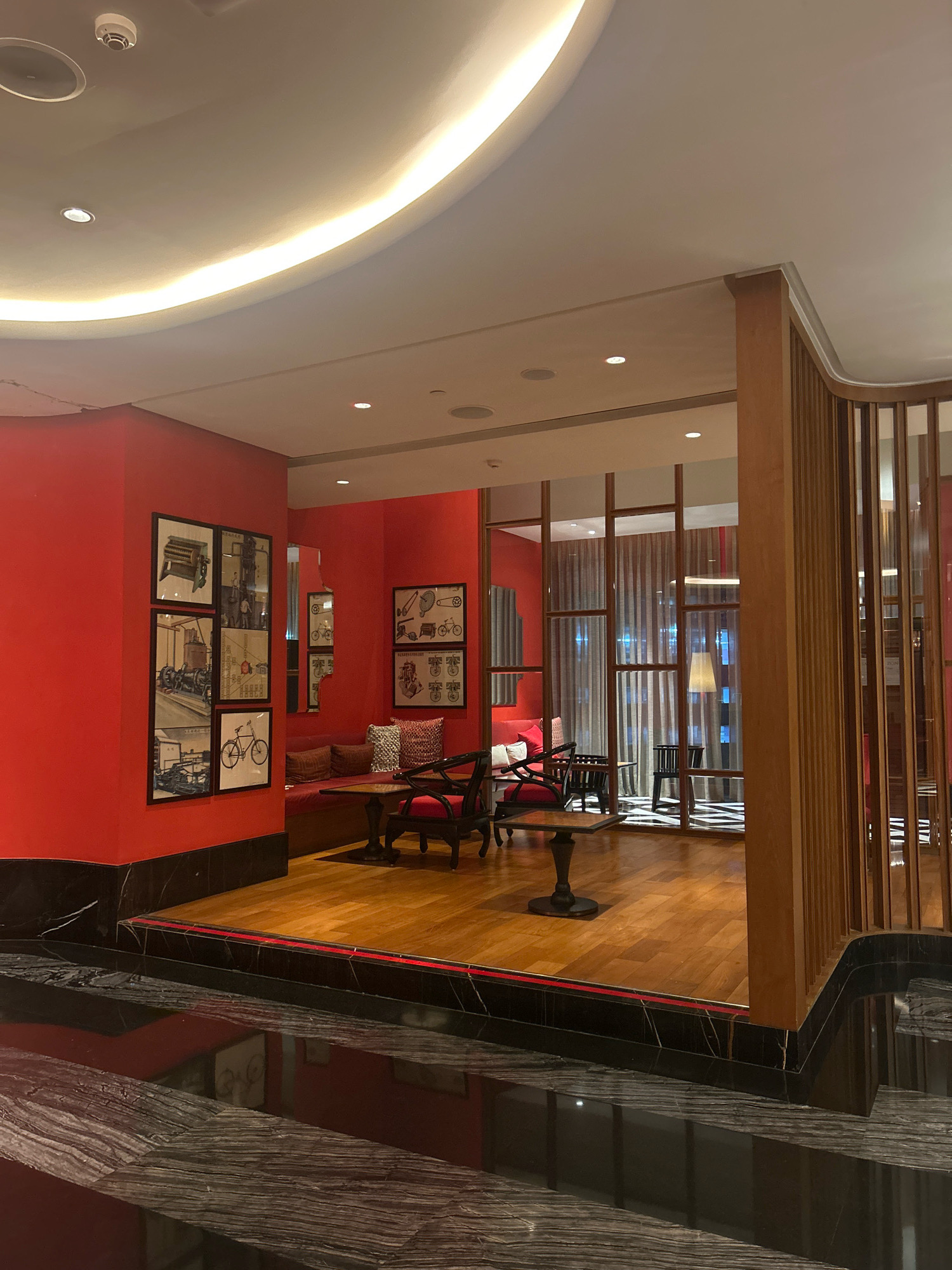 The image shows an interior scene of a lounge or waiting area, likely a hotel lobby or restaurant, in Hyderabad, India. The space features a vibrant red wall with framed black and white prints depicting vintage bicycles and industrial machinery. A sunken seating area with red cushions and patterned throw pillows is visible, along with a dark wooden table and chairs. The flooring is a combination of polished dark marble and warm-toned wood. A decorative wooden screen with vertical slats partitions off a section of the area, revealing more seating and curtains in the background. The ceiling is modern and curved, with integrated lighting and a smoke detector. The overall ambiance suggests a stylish and contemporary design with a touch of retro flair.