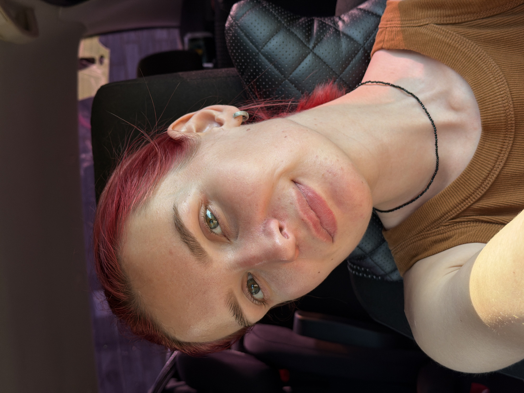 The image is a close-up selfie of a young woman with red hair and green eyes, taken inside a car. She is wearing a brown ribbed tank top and a thin black beaded necklace. She has a silver hoop earring in her left ear. The background shows the interior of a car, with dark leather seats. The lighting suggests it is daytime, with sunlight coming from the left. The woman is smiling slightly and looking towards the camera. There is no indication of the city of Chaikovsky, Russia, or any specific urban environment.
