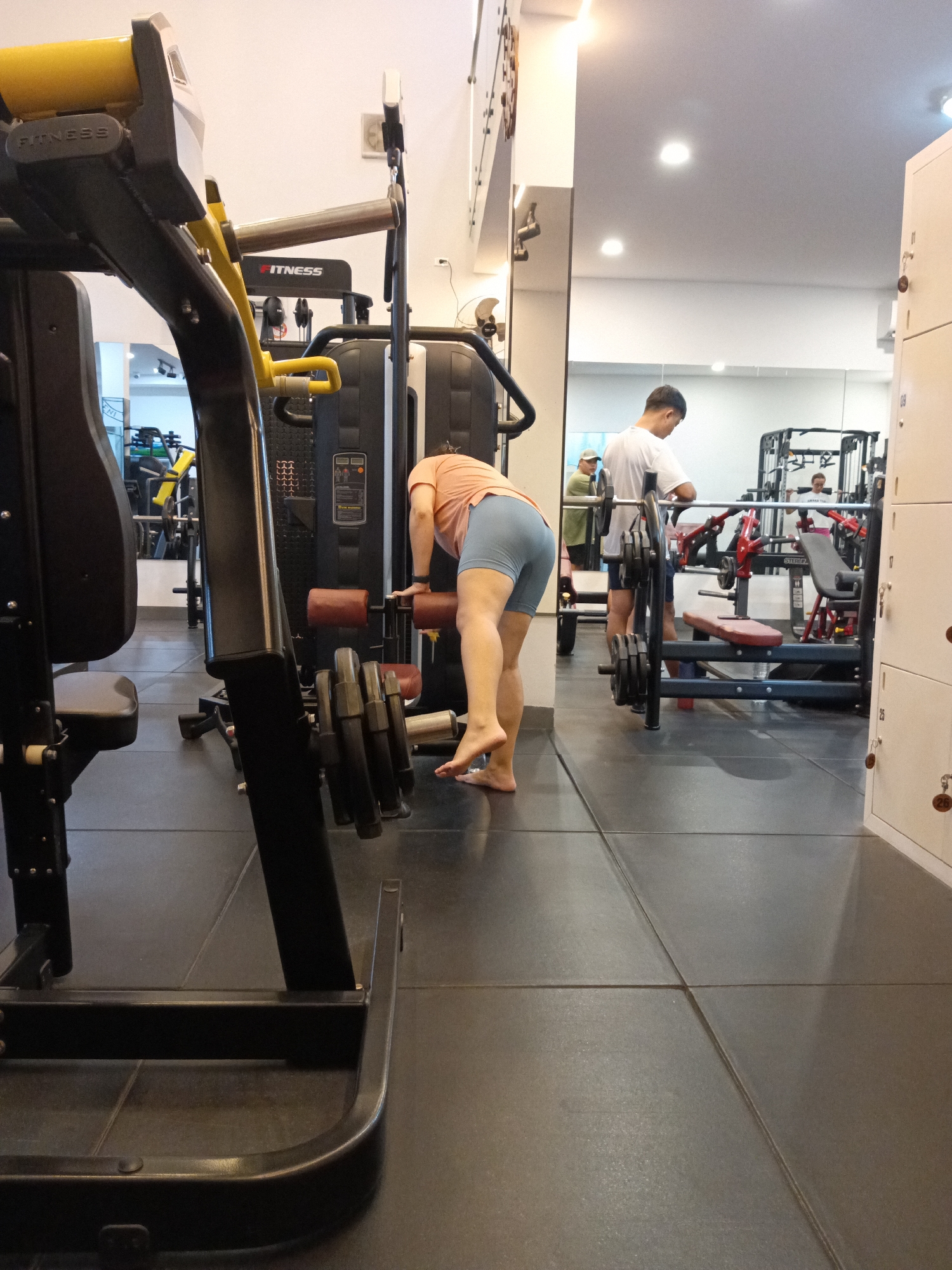 This image depicts the interior of a gym in Phu Thuy Ward, Vietnam. In the foreground, a person wearing an orange t-shirt and blue shorts is bending over, facing away from the camera, and appears to be adjusting a weight machine. Their feet are bare. To the left, a large piece of gym equipment with black and yellow accents is partially visible. In the background, a man in a white t-shirt and blue shorts stands near a weightlifting bench and barbell. Another person, wearing a green cap and a lighter colored shirt, is visible behind him. The gym has tiled flooring and a mirrored wall in the background, reflecting more gym equipment. Lockers are visible on the right side of the image.