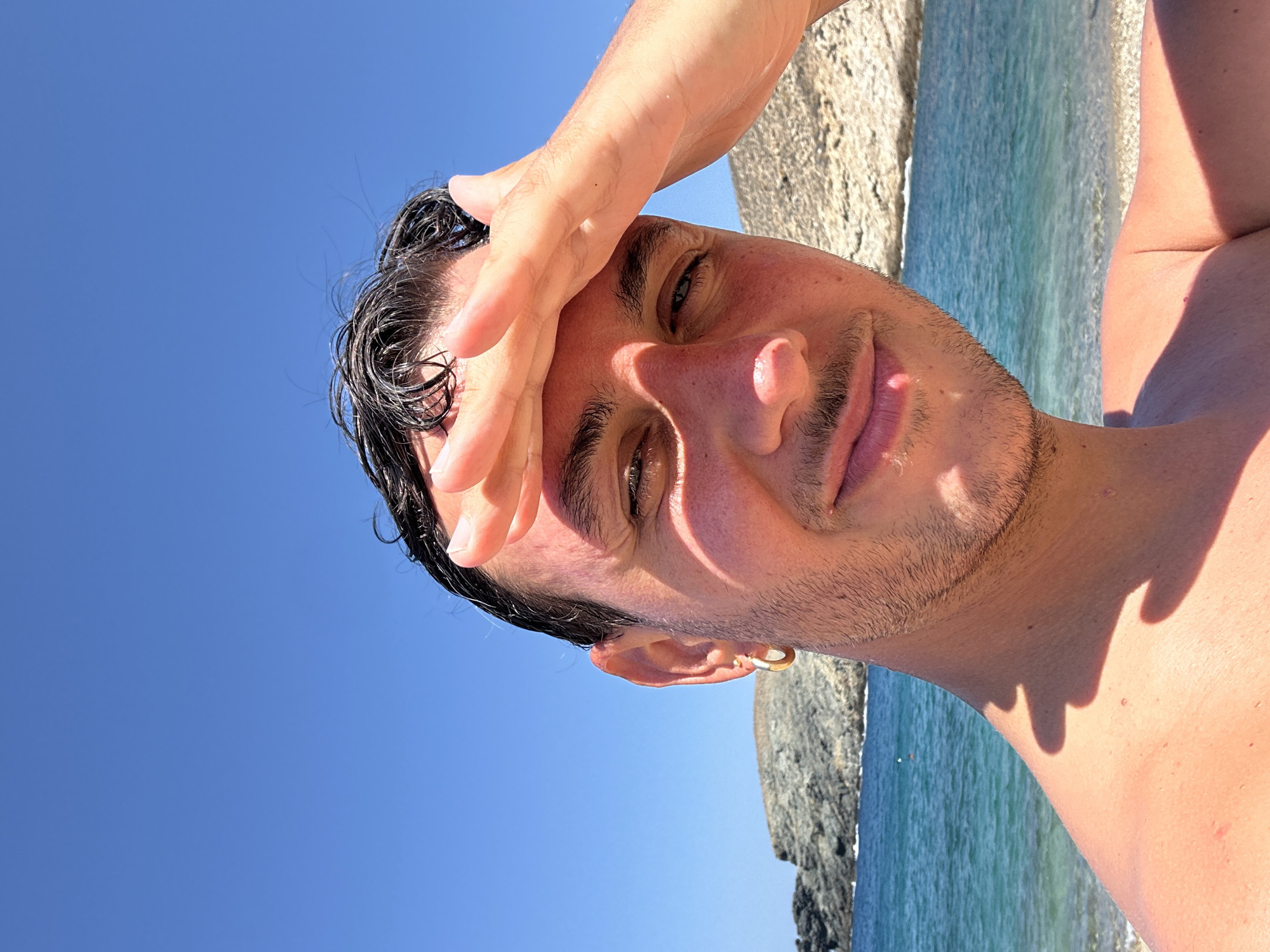 A close-up, selfie-style medium shot captures a young man at a beach in Ano Mera, Greece. He is positioned in the foreground, looking towards the camera with a slight smile, his right hand raised to shield his eyes from the sun. His dark, wet hair is slicked back, and he has a faint mustache and stubble. He is wearing a gold hoop earring in his left ear. The background features the clear blue sea meeting a sandy beach, with rocky hills rising behind the water. The sky is a bright, cloudless blue. The lighting suggests a sunny day.
