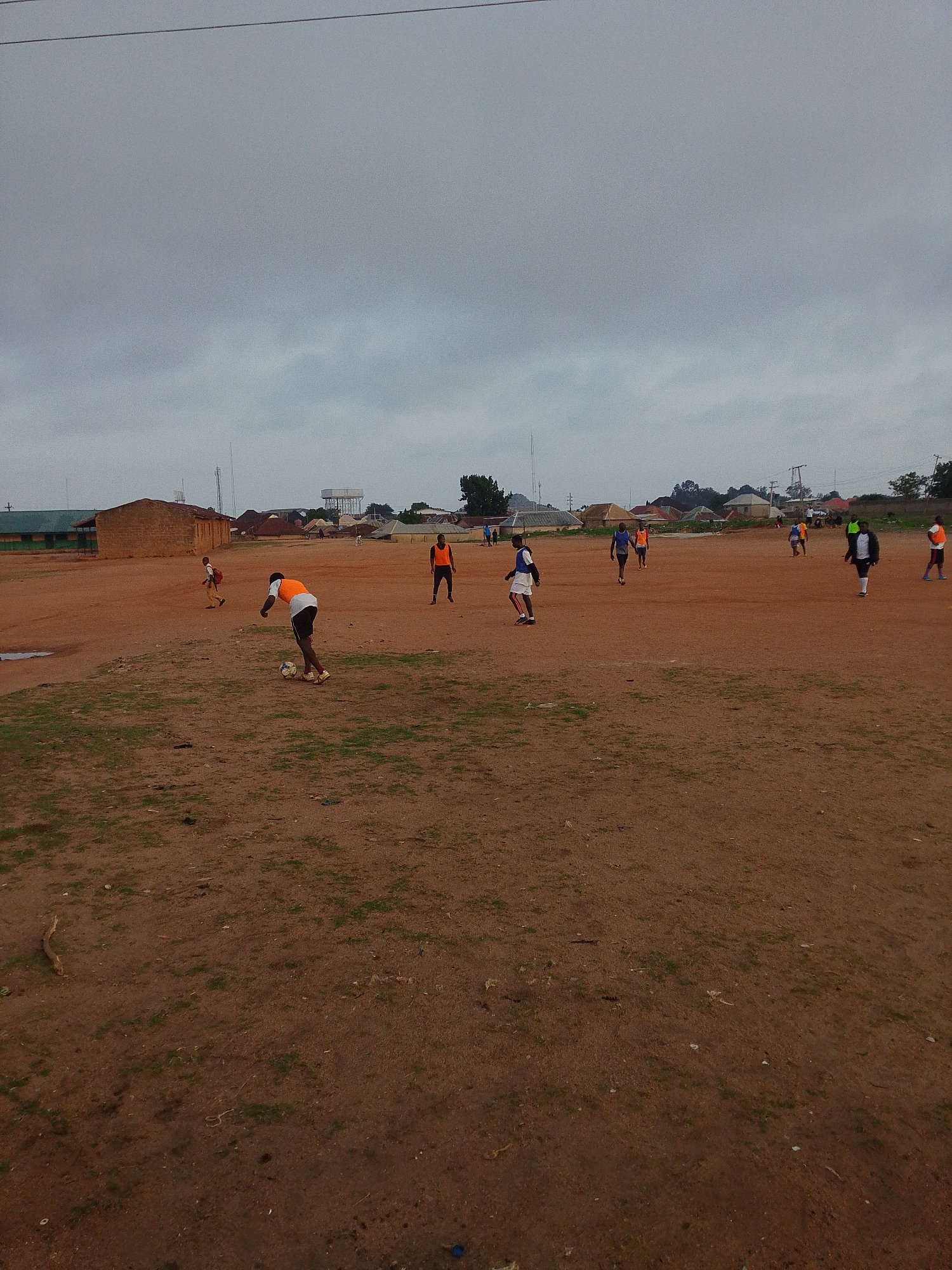 The image shows a soccer match in progress on a red dirt field. Several players are scattered across the field, some actively engaged with the ball, while others are jogging or standing. The players are wearing a mix of white, blue, orange, and black jerseys. In the background, there are low-rise buildings, and a water tower. The sky is overcast with heavy grey clouds. The location is Bukuru, Nigeria, based on visual characteristics of architecture and environment.