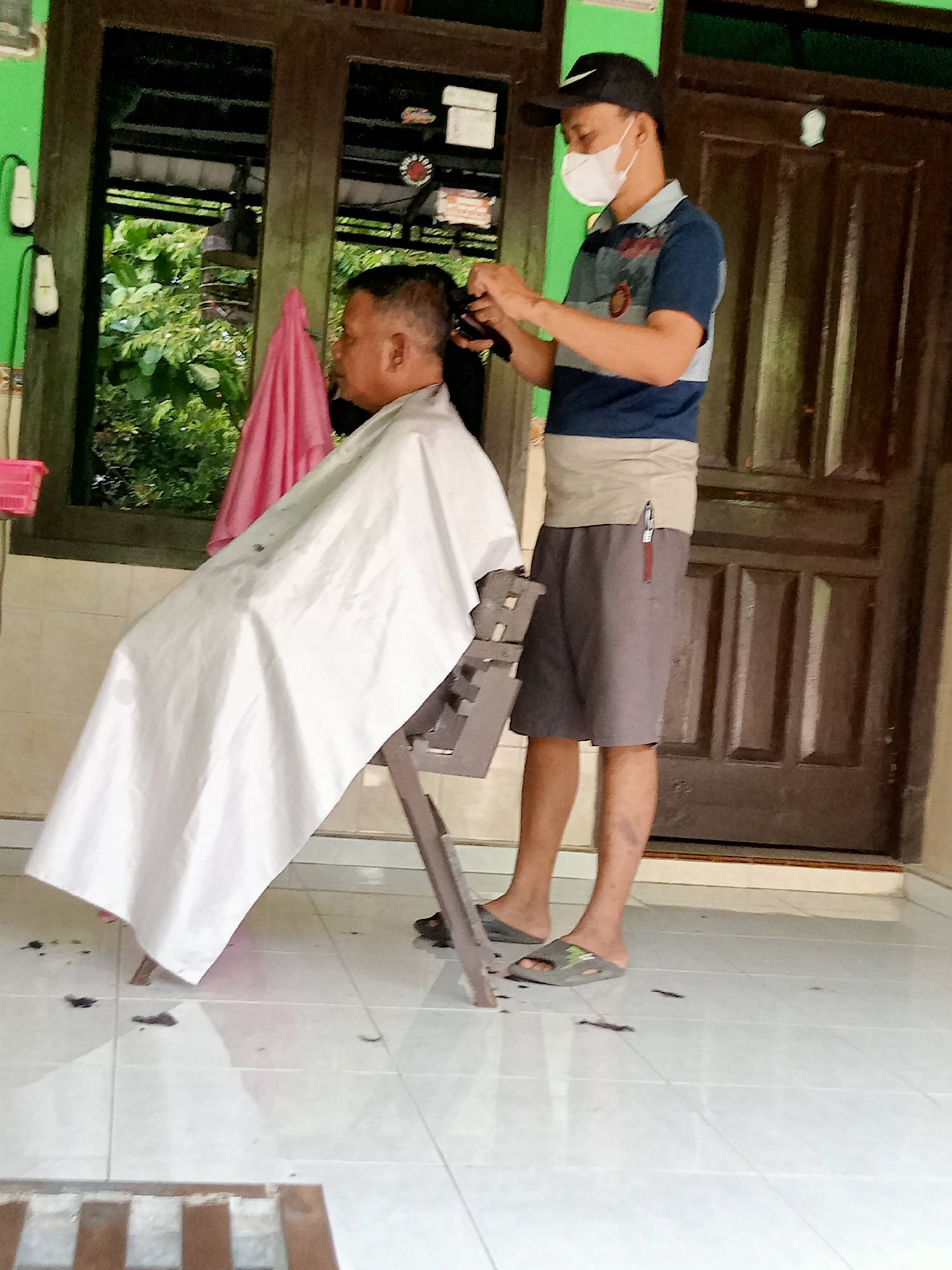 In Majan, Indonesia, two individuals are engaged in a haircut outdoors. A man, seated and draped in a white cape, is receiving a haircut from another man who is standing and holding clippers. The barber is wearing a blue and beige striped t-shirt, grey shorts, a black baseball cap, and a white face mask. The scene appears to be in front of a residential building with a dark wooden door and green walls. Green foliage is visible in the background through a window. Clippings of hair are scattered on the light-colored tiled floor.