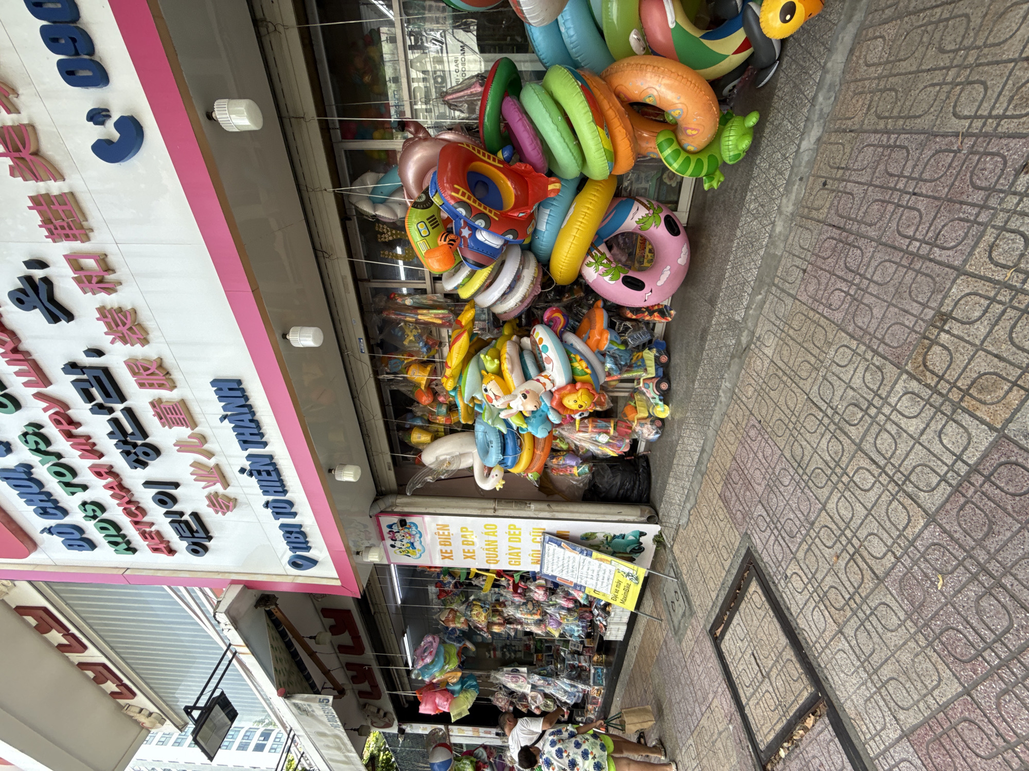 This is a street-level, eye-level, medium shot of a toy store in Nha Trang, Vietnam, located at 1181 Tô Hiến Thành street. The store front displays a variety of inflatable toys, including inner tubes, pool floats, and character-themed inflatables. Colorful balloons and other packaged toys are also visible inside the store. A sign in Vietnamese advertising "Xe điện, Xe đạp, Quần áo, Giày dép, Nôi cũi" (Electric vehicles, Bicycles, Clothing, Shoes, Baby cribs) is posted outside the store. Two individuals are present: a woman in a green patterned shirt and shorts is walking away from the store with a shopping bag, and a man wearing a white t-shirt is browsing items inside. The sidewalk in front of the store is paved with patterned tiles, and a manhole cover is visible. The store's facade features prominent signage with the store's name and offerings in multiple languages, including English and Korean.
