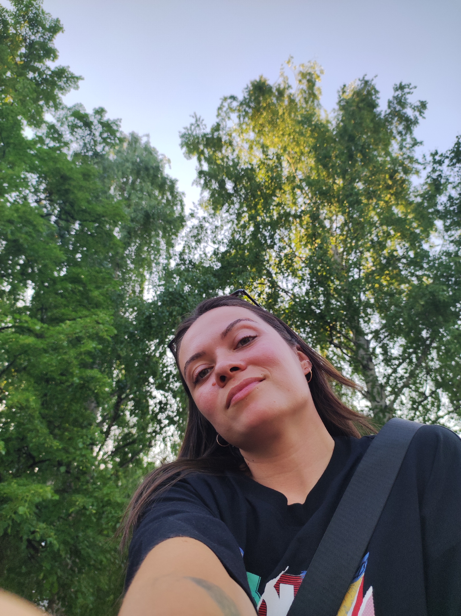 A selfie of a woman with fair skin and brown hair, taken from a low angle, with green trees and a pale blue sky in the background. The woman is wearing a black t-shirt and a gold hoop earring in her left ear. A black strap is slung across her chest. A pair of sunglasses are tucked into her hair. The location is possibly a park or wooded area in the city of Chaikovsky, Russia, based on the presence of trees, though the specific location is not identifiable from this image. No specific actions or interactions are visible. The environment appears to be outdoors in daylight.