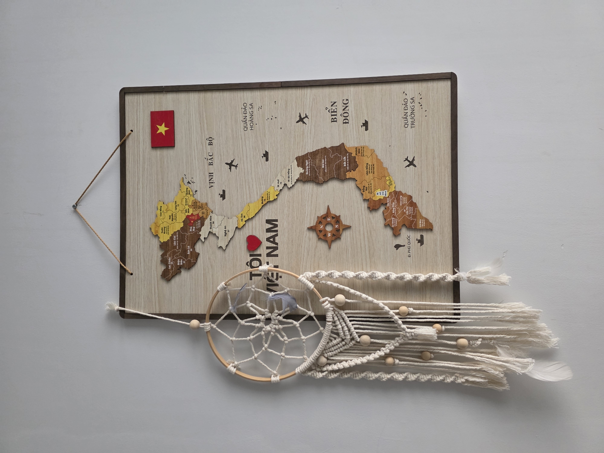 This image depicts a wooden map of Vietnam, complete with regional demarcations and place names printed on the surface. The map is framed in a dark brown wood, and a small Vietnamese flag is attached to the upper left corner. Below the map, a dreamcatcher hangs, adding a decorative touch. The phrase "TÔI VIET NAM" is printed on the map near the middle. The map is likely displayed in a room in Mui Ne, Vietnam.